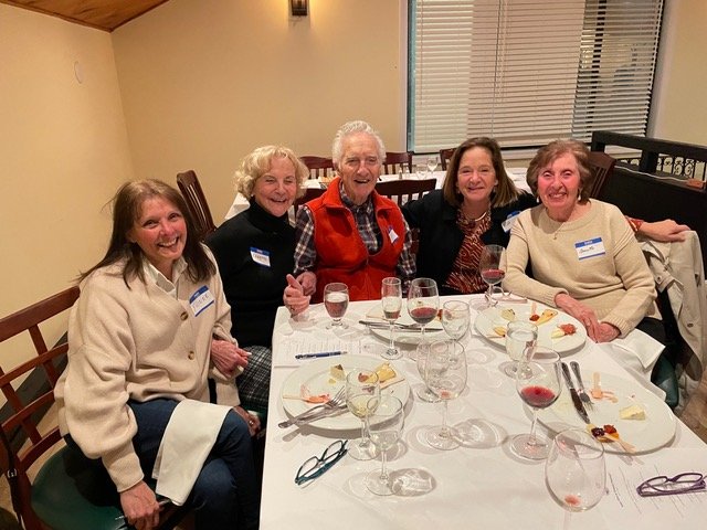 A group of five seniors sitting at a restaurant table, smiling. They are dressed in casual, warm clothing. The table has glasses, a small plate of food, and name tags are visible on their clothing.