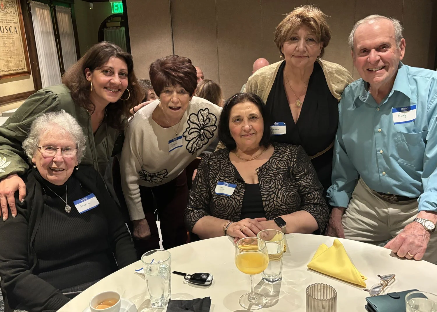 Group of seven adults gathered around a table at a social event, smiling for the camera, with drinks, napkins, and utensils on the table.