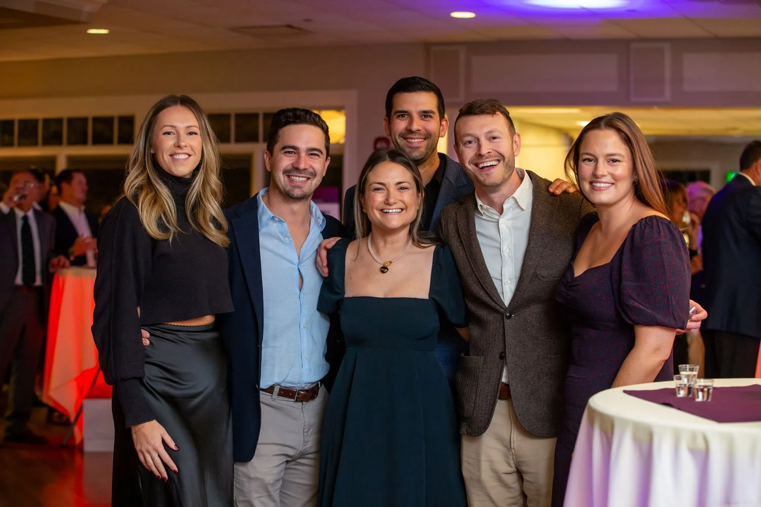 Group of six young adults smiling at a social event, standing close together indoors with tables and other attendees in the background.