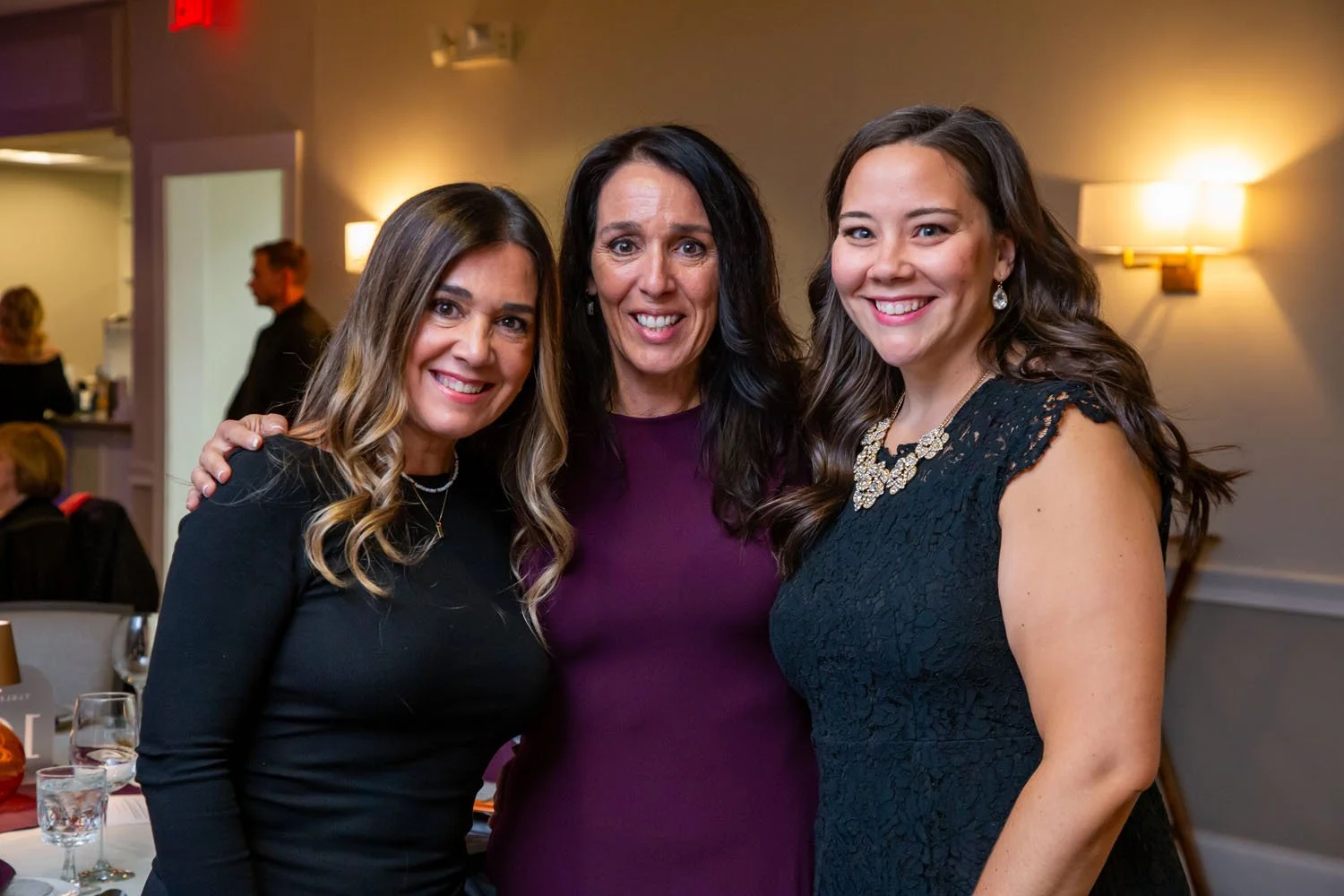Three women at a social event, dressed nicely, smiling and posing together indoors with warm lighting.