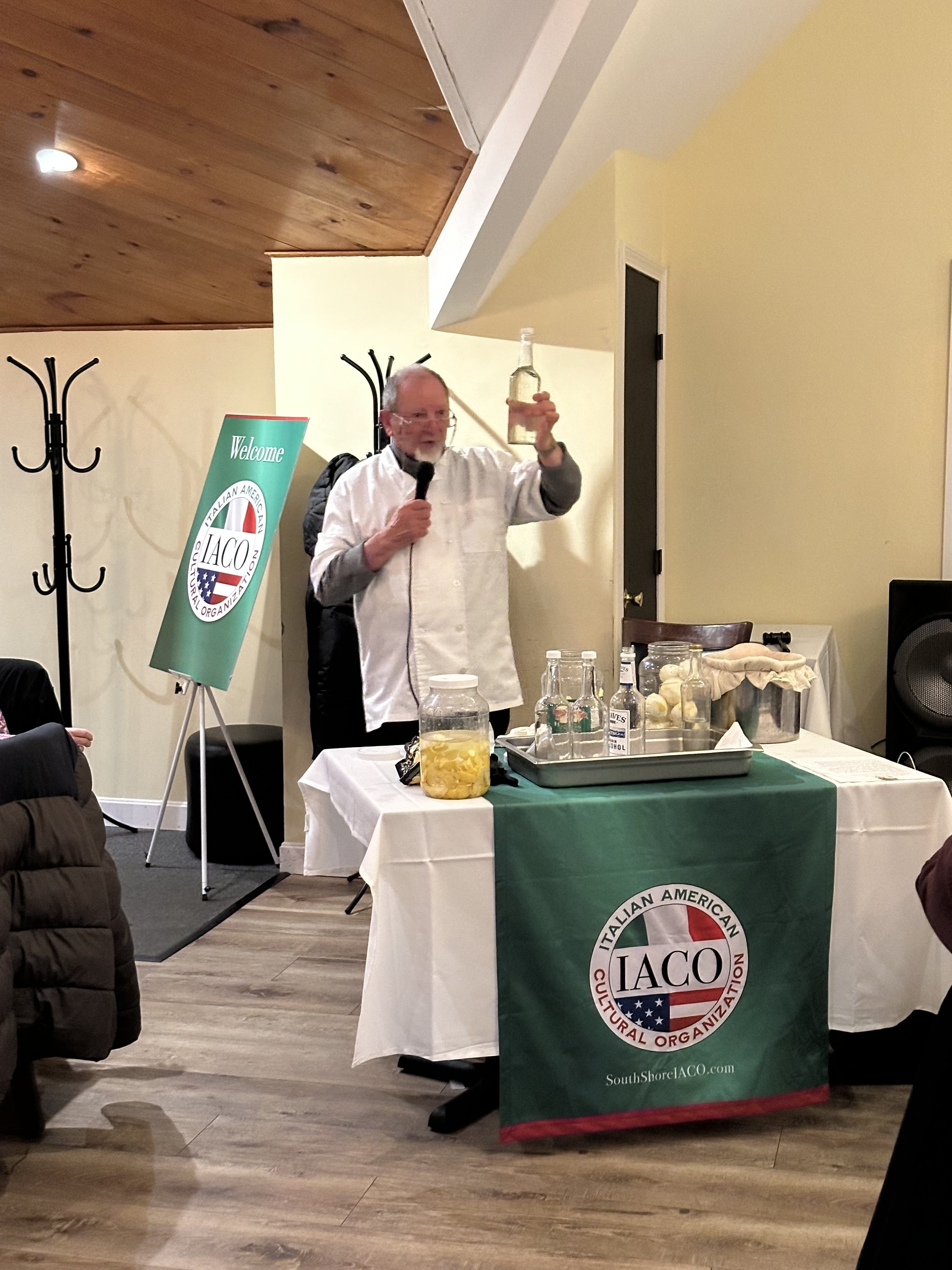 A person holding a clear bottle speaks into a microphone at a presentation for the Italian American Cultural Organization. A table with jars of lemons and bottles is covered with a tablecloth featuring the organization's logo and website. A display b