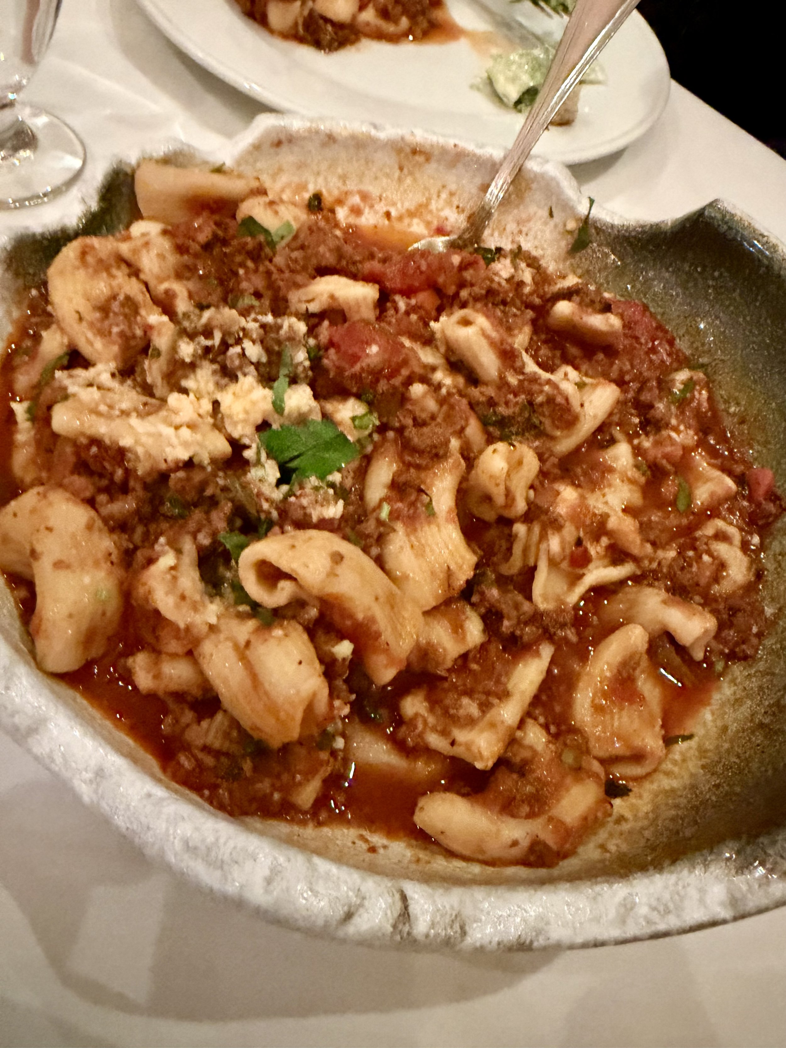 Large bowl of pasta with meat sauce, garnished with parsley, on a white table. In the background, there is a glass of water and a plate with salad.