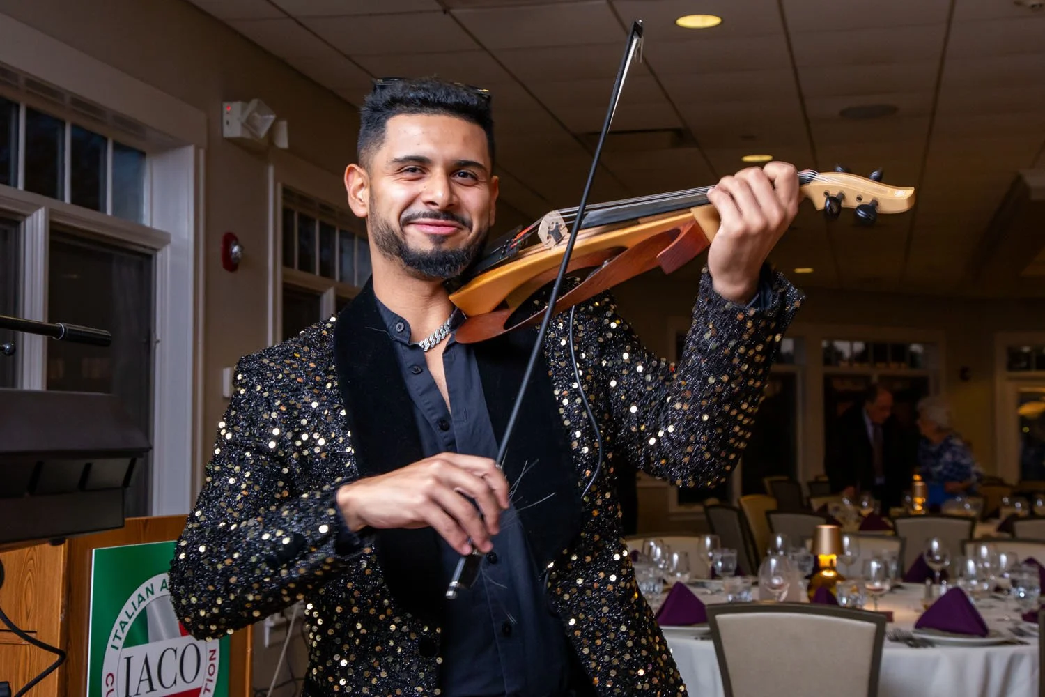 A man with dark hair and a goatee, wearing a black sequined jacket and a chain necklace, is smiling and playing a violin at an indoor event with tables, glasses, and purple napkins.
