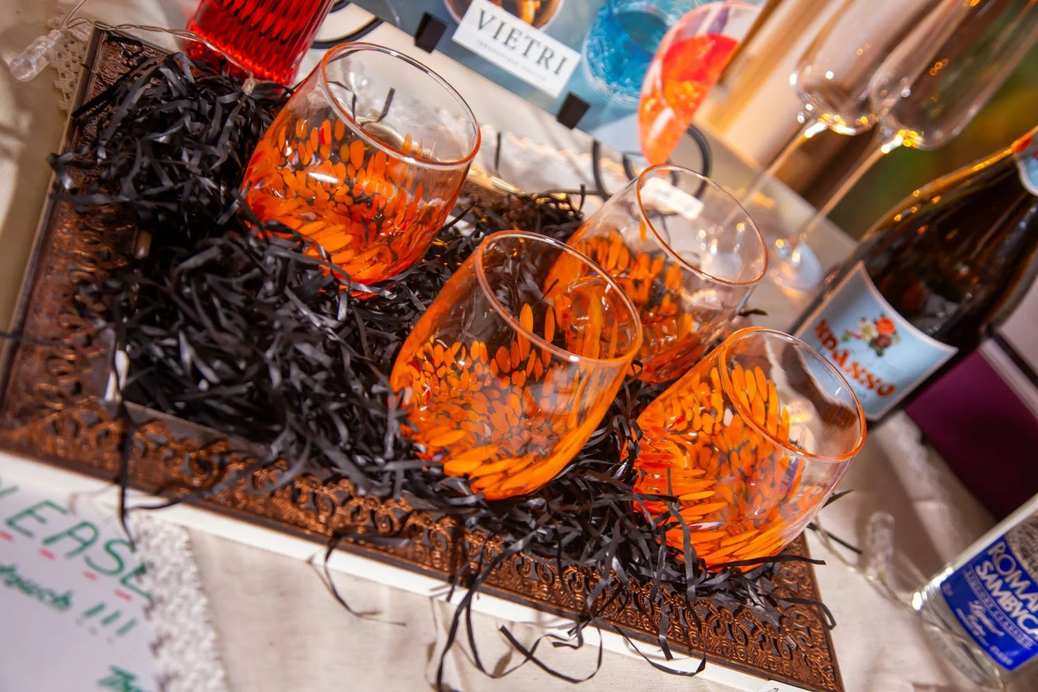 Four glasses of orange-colored drink with ice on a decorative tray filled with black shredded paper, surrounded by bottles and other items on a table.