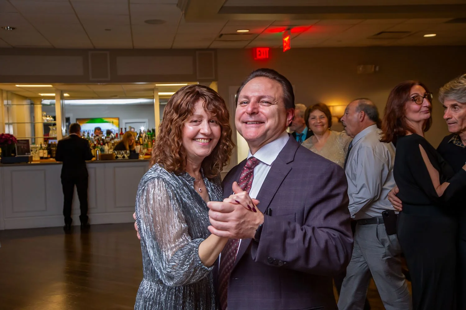 A group of people dancing at a social event in an indoor venue, with a bar in the background.