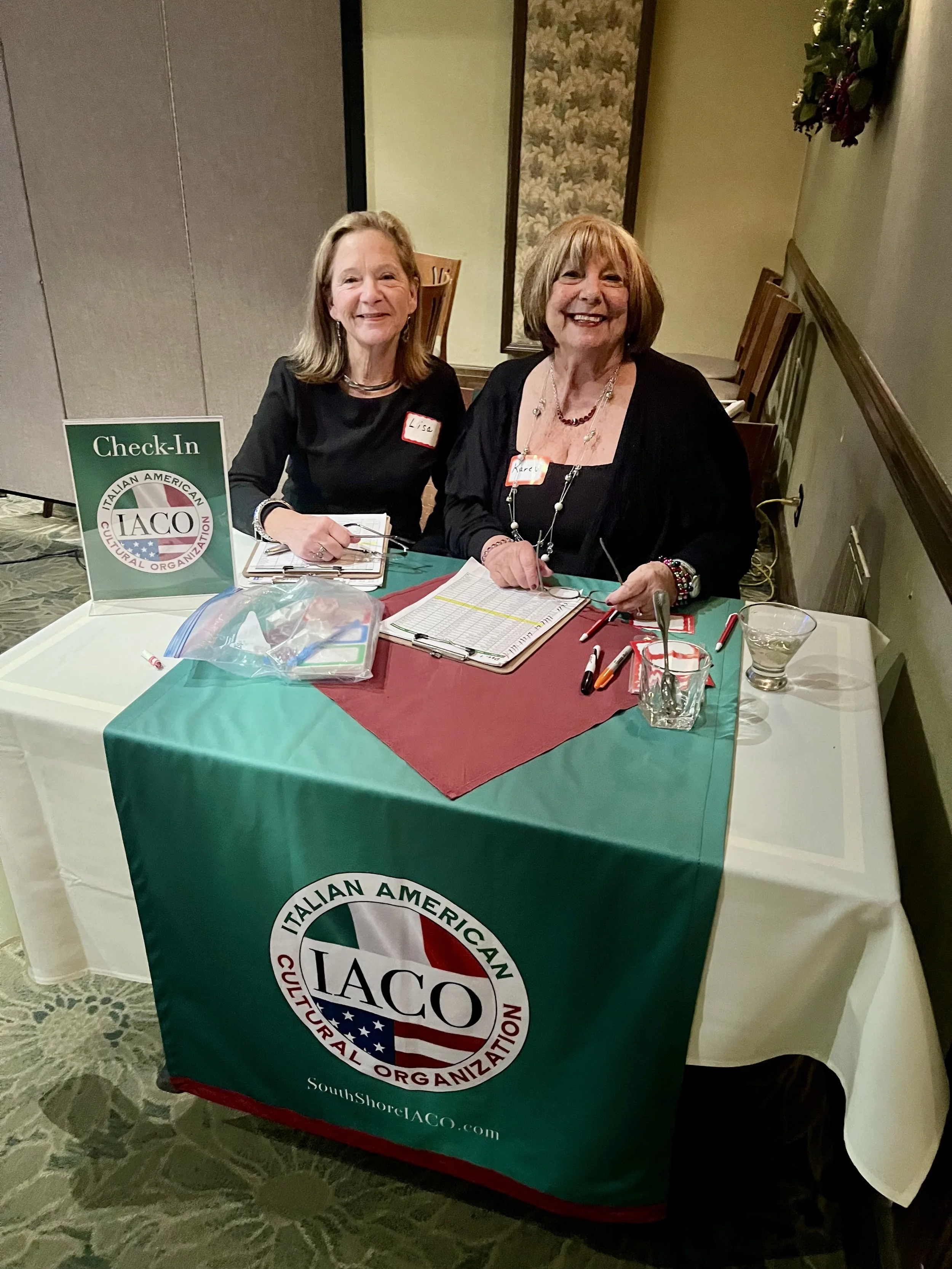 Two women sitting at a table with an IACO banner at an event, smiling at the camera. The table has clipboards, pens, and drinks on it.
