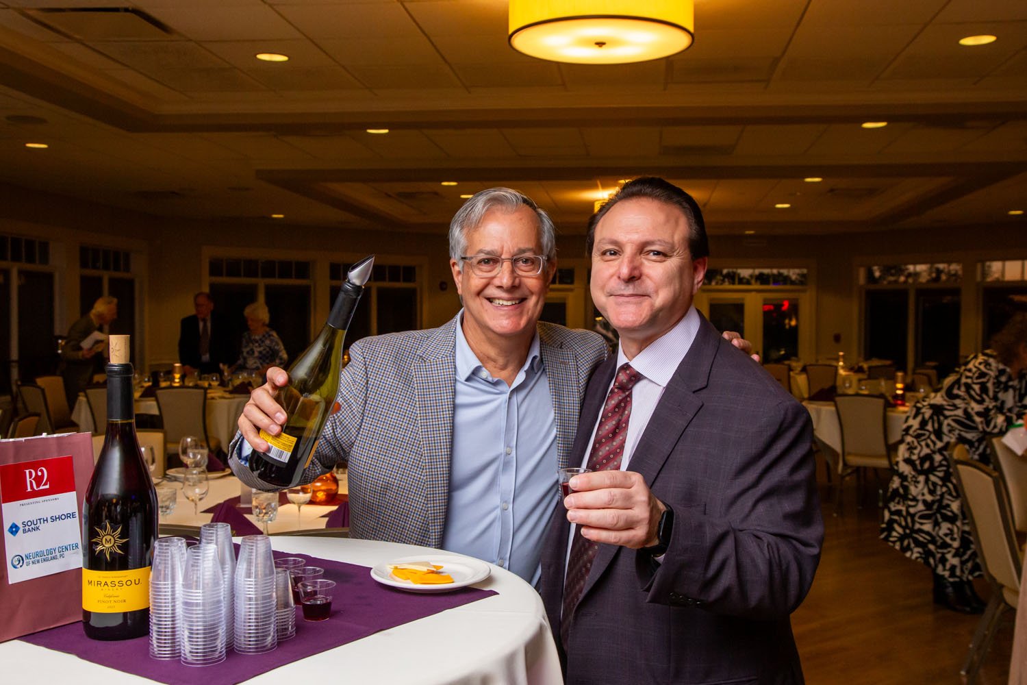 Two men in formal attire smiling and raising glasses at a social event or dinner party, with wine bottles and plastic cups on the table.