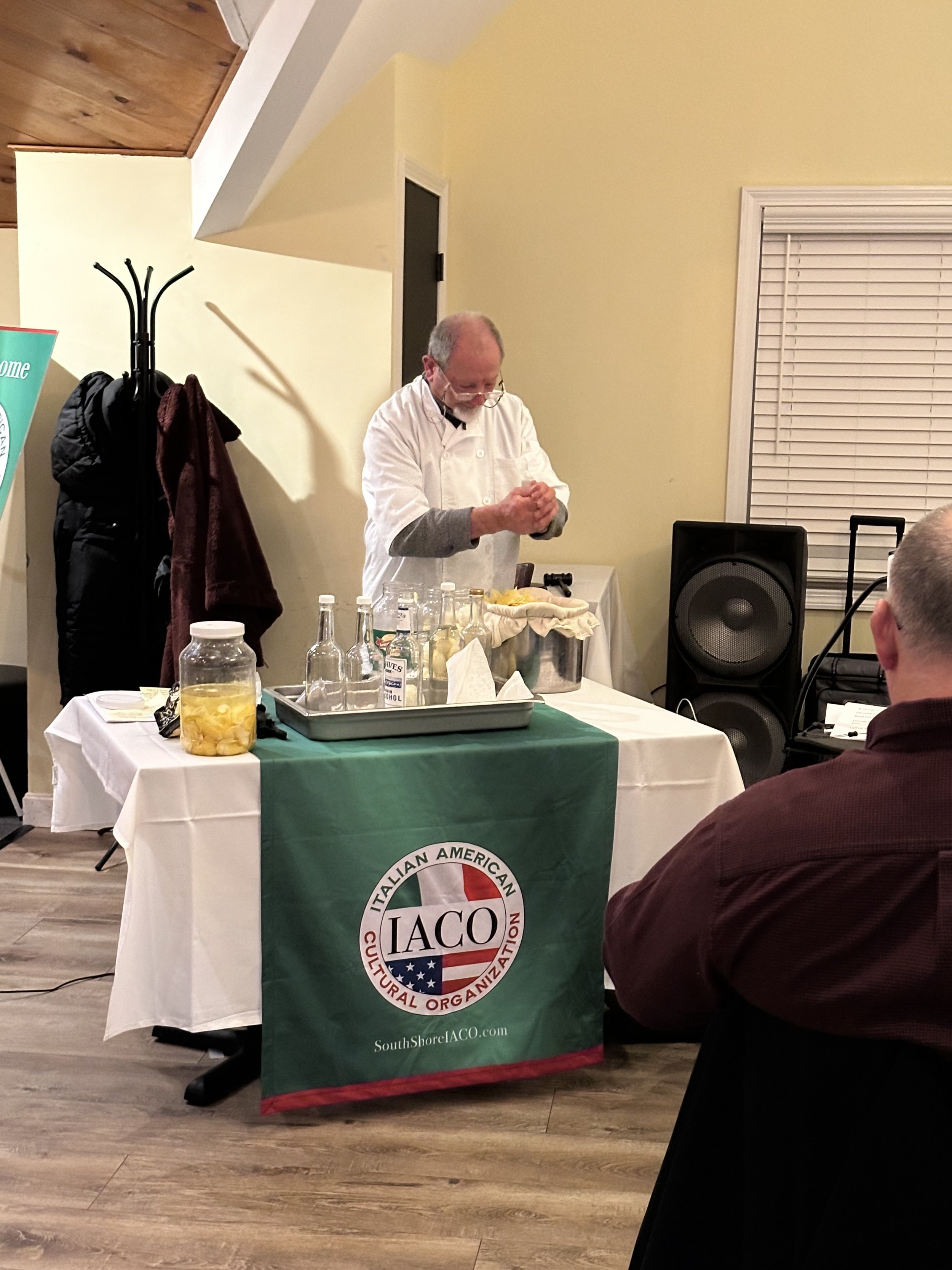 Man demonstrating cheese making at a table with bottles and jars, covered with a cloth featuring the IACO logo and name, inside a room with people watching.