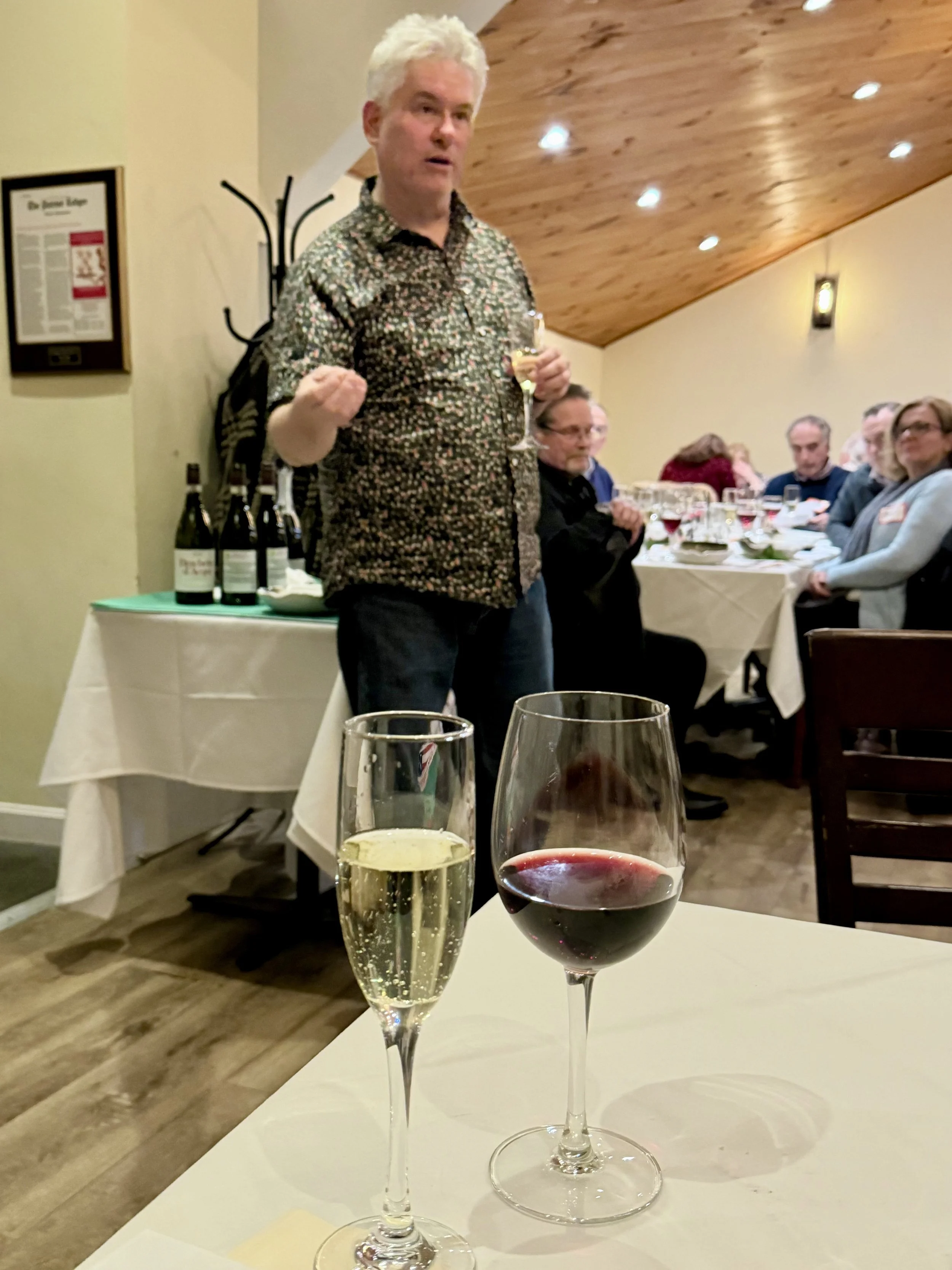 A man with white hair speaking at a dinner event, holding a wine glass. There are two glasses of wine, one with white wine and the other with red wine, on the table in the foreground. Several people are seated at tables behind him in a warmly lit roo