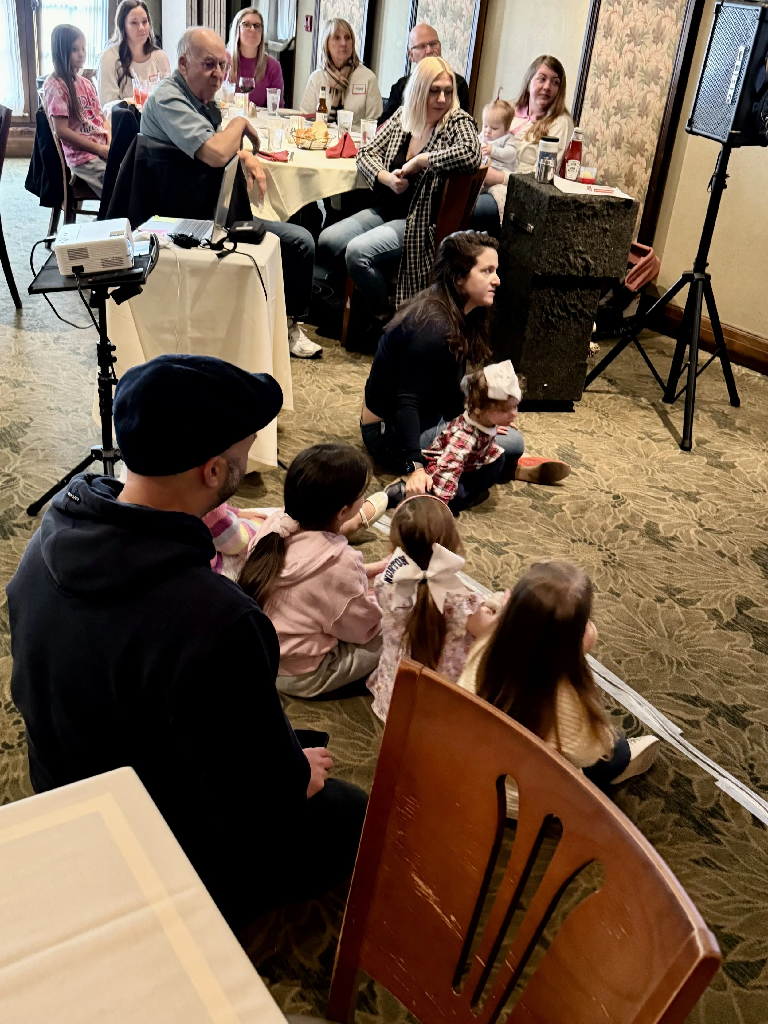 People sitting on the floor and at tables in a restaurant or banquet hall. Children in the front, watching an event, with adults seated behind them. Equipment such as a projector and speaker are visible, indicating a presentation or performance.