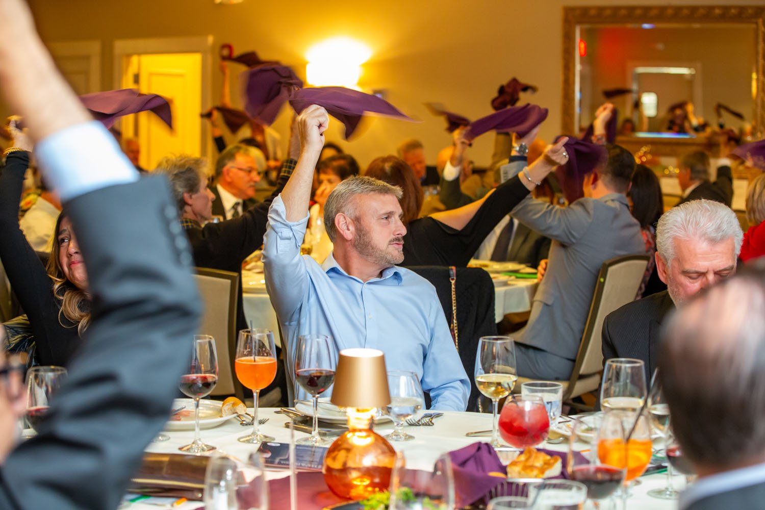 People seated at a banquet table during a formal event, waving purple napkins in the air.
