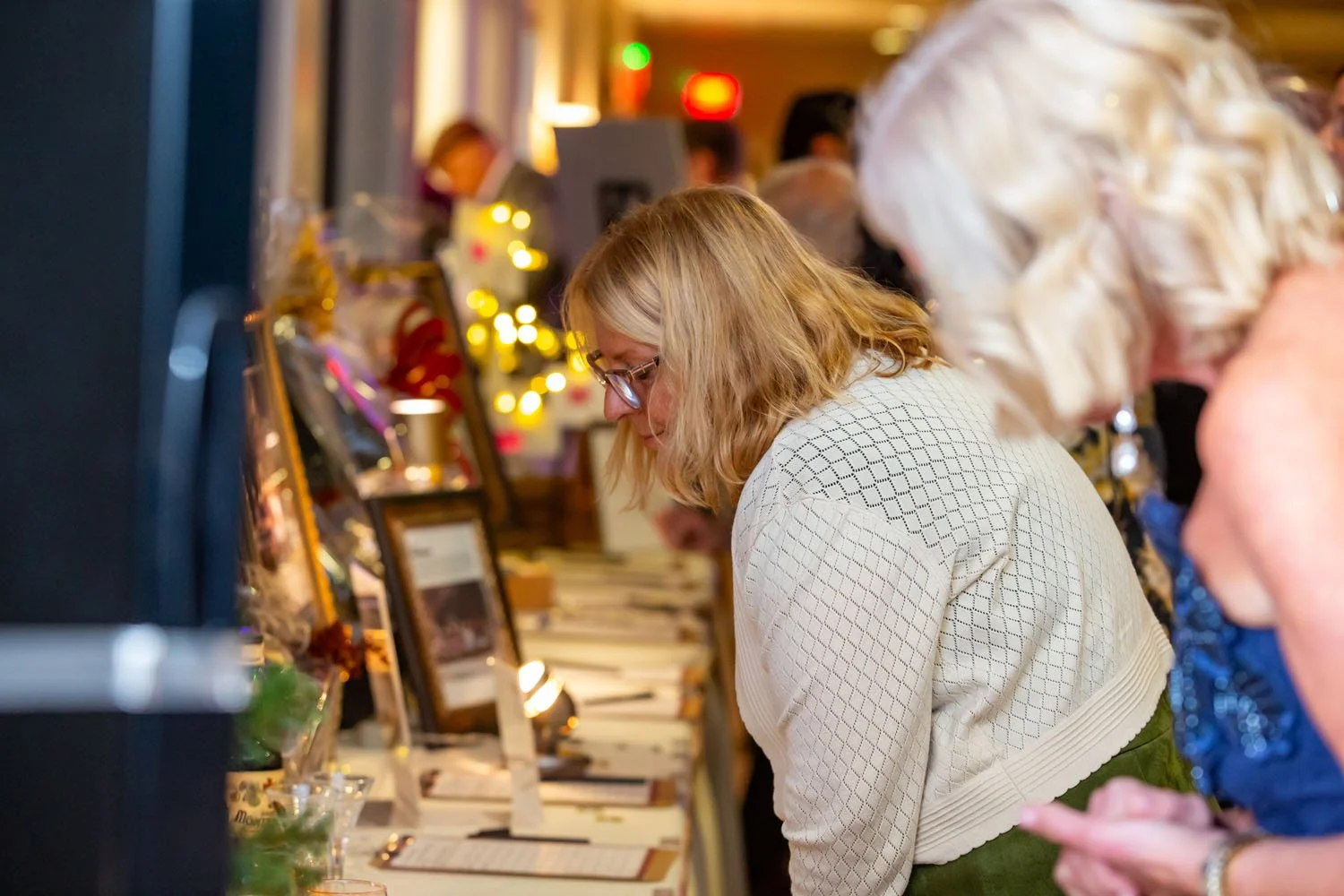 Woman with blonde hair and glasses looking at items on display at an indoor event with holiday decoration in the background.