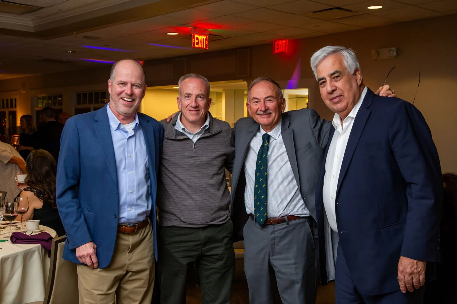 Four men standing together at a social gathering, smiling at the camera, with a dinner event in the background.