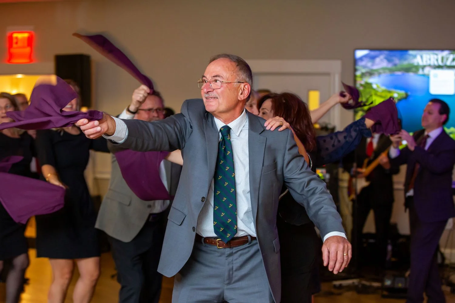 Man in a gray suit dancing with purple napkins at a celebration event with other people dancing and playing instruments in the background.