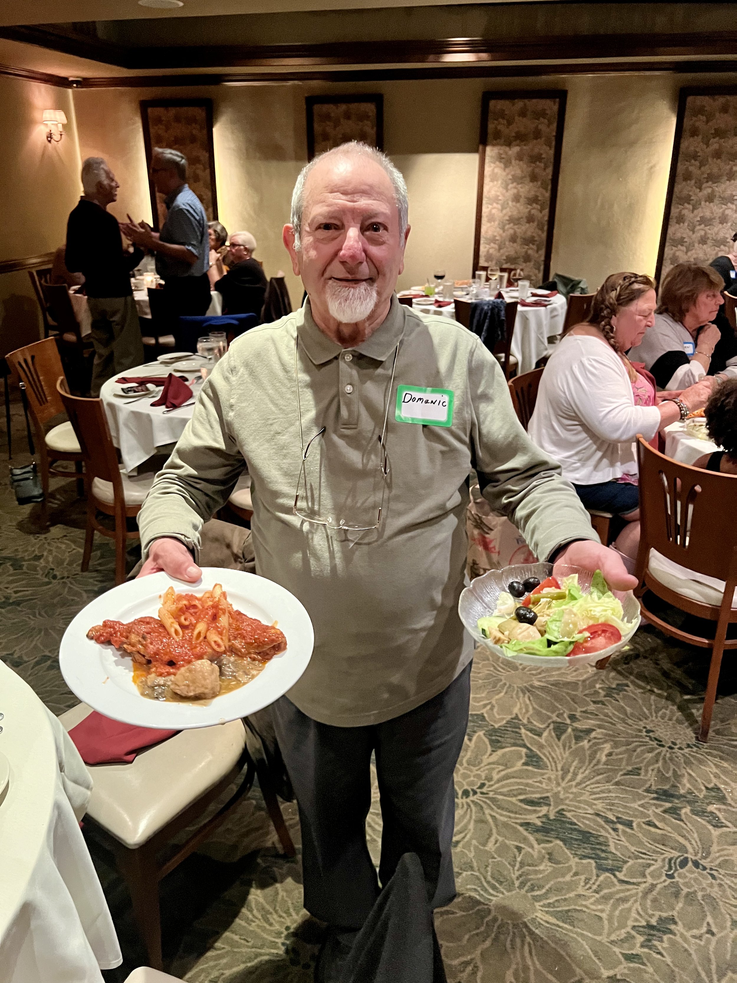 A man in a restaurant holding a plate of pasta with tomato sauce and a salad bowl. The background shows other people seated at dining tables.