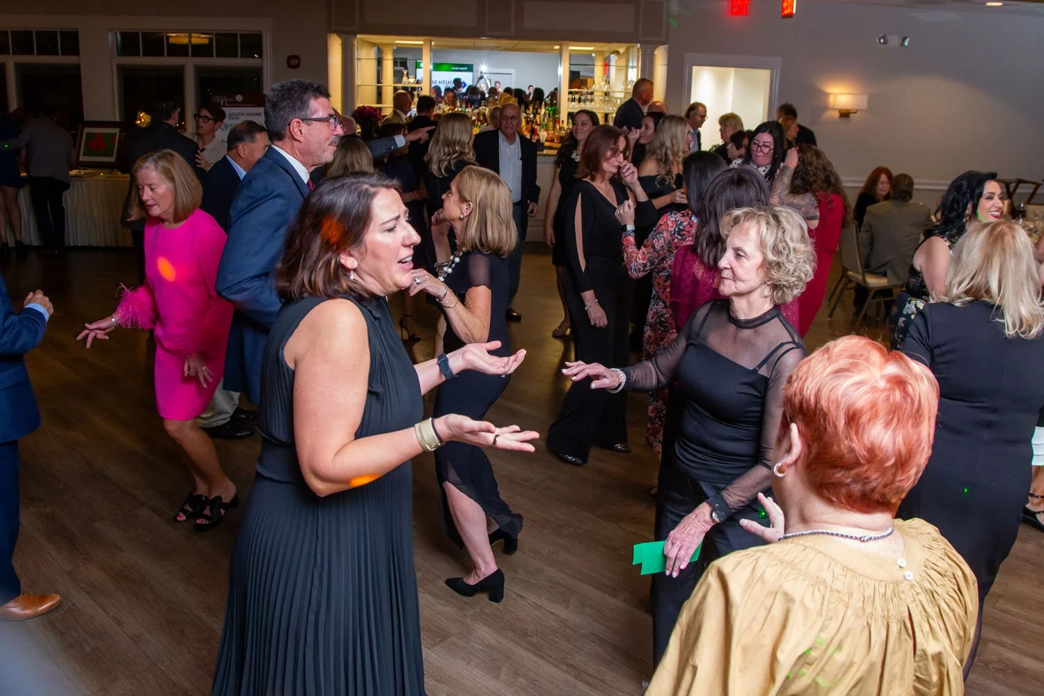 People dancing and socializing at an indoor party or event, with a bar in the background and various guests dressed in formal to semi-formal attire.