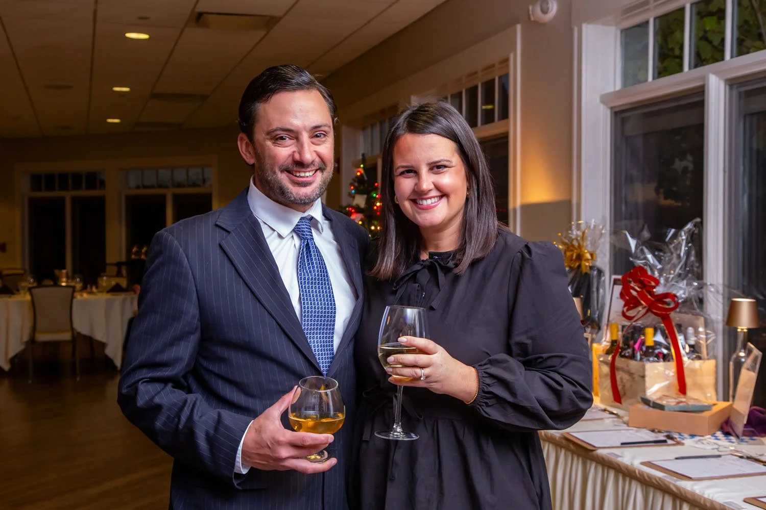 A man and woman in formal attire smiling at the camera, holding wine glasses, at a holiday celebration in a decorated banquet hall.