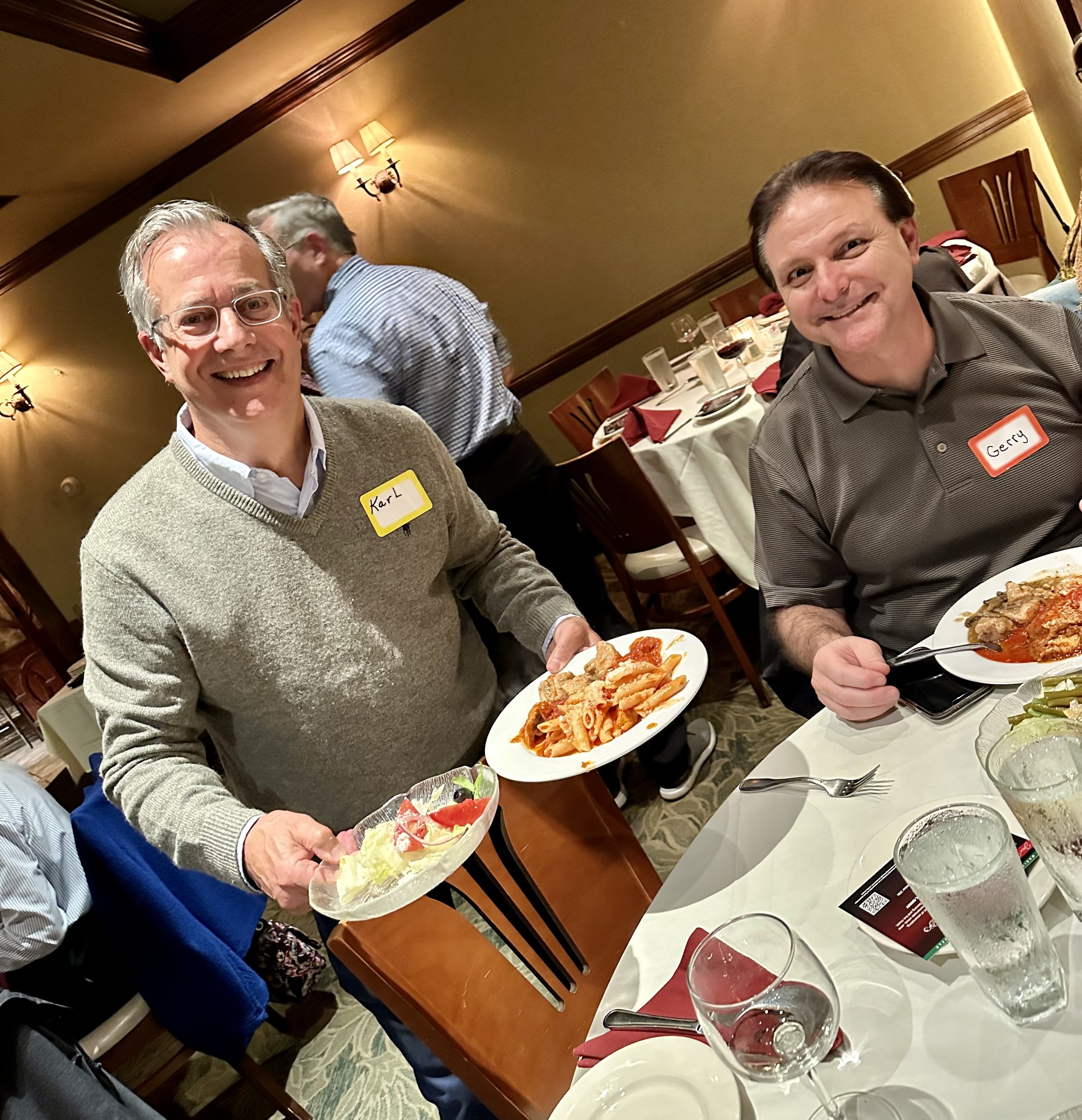 Two men at a restaurant table with plates of food, one with pasta and salad, the other with a meat dish, smiling at the camera.