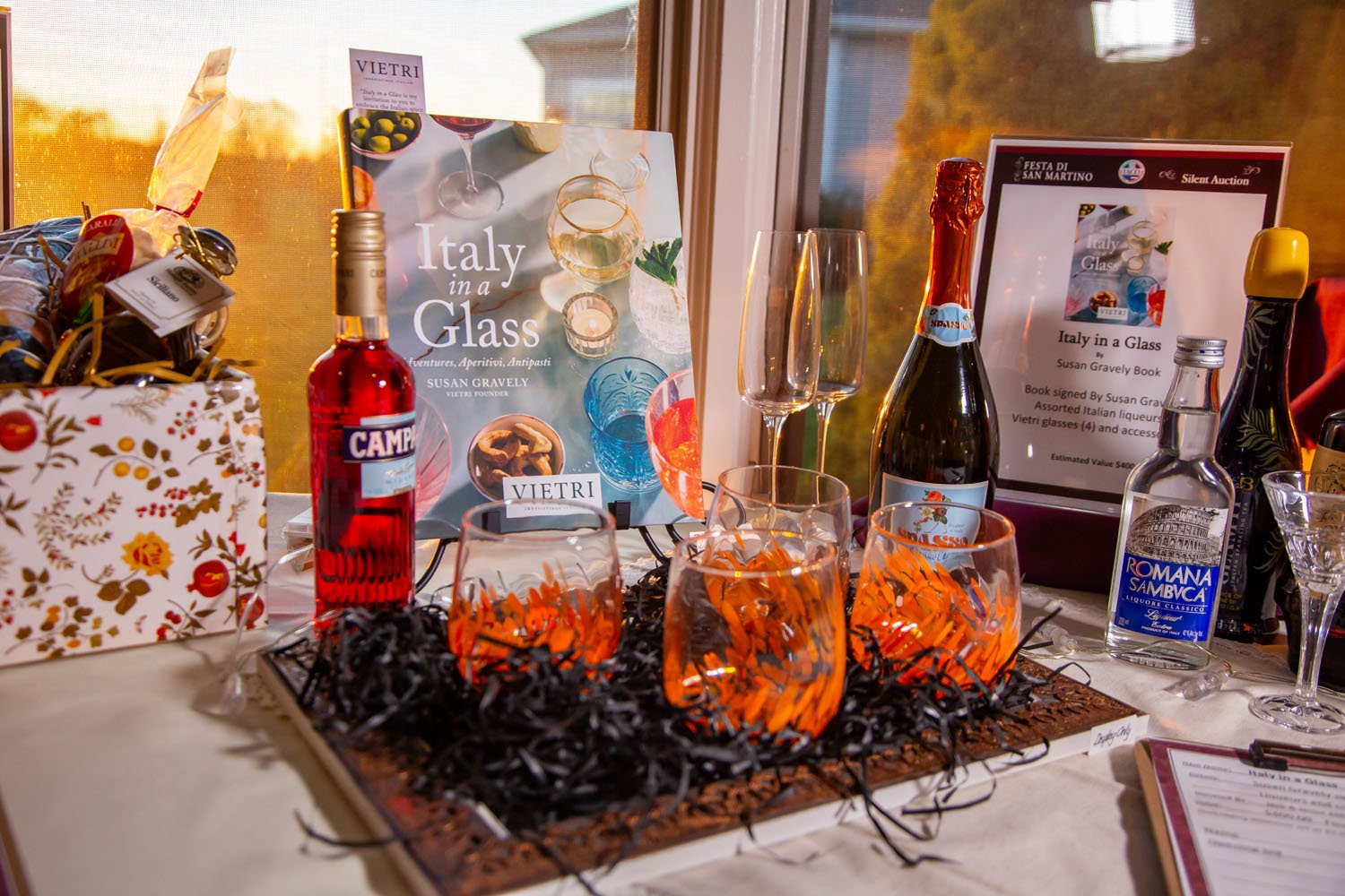 Display of Italian wine and liquor bottles, glasses, and related promotional materials on a table at an event, with a window showing trees and sunset in the background.