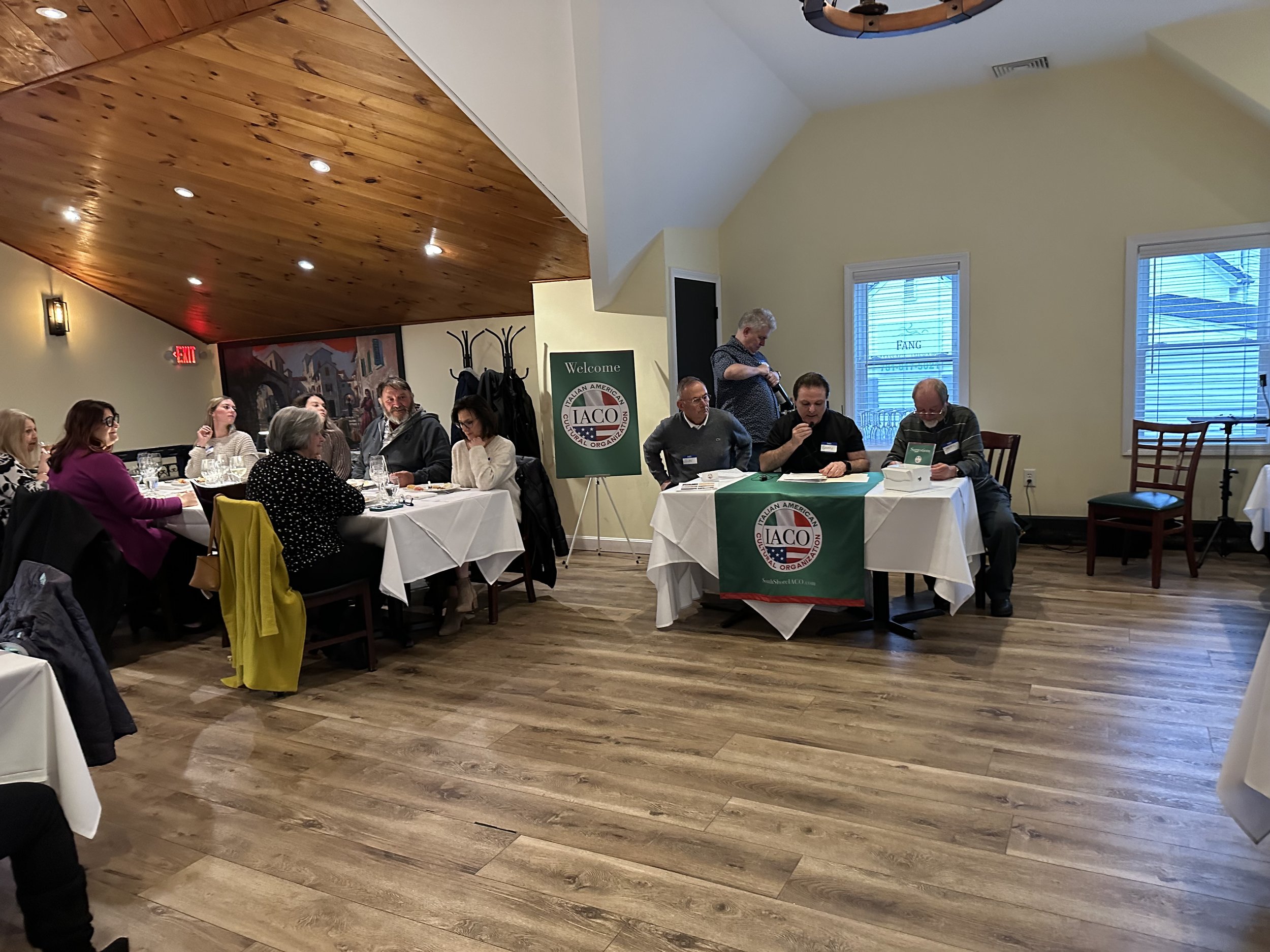 A group of people sitting at tables in a restaurant event room with a sign displaying 'IACO' (Italian American Club of the Carolinas) in the background. Some are conversing and others appear to be conducting a formal discussion or meeting.
