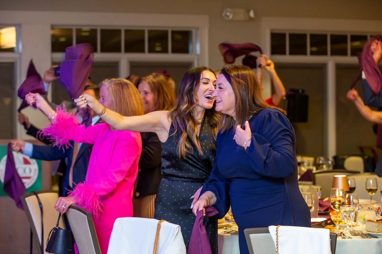 Women celebrating at a party, waving purple cloths, smiling and enjoying themselves, seated at tables with glasses and tableware.