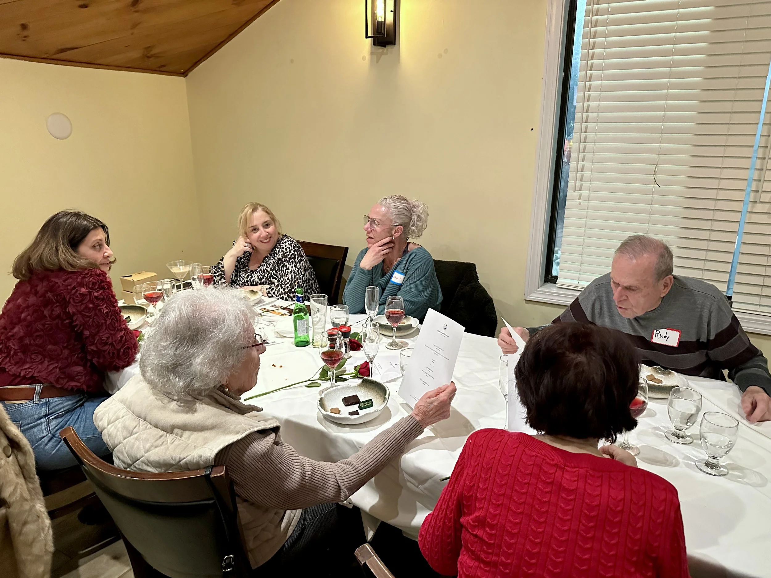 A group of people sitting around a decorated dinner table with plates, glasses, and drinks, engaged in conversation, in a warmly lit room with a window and blinds.