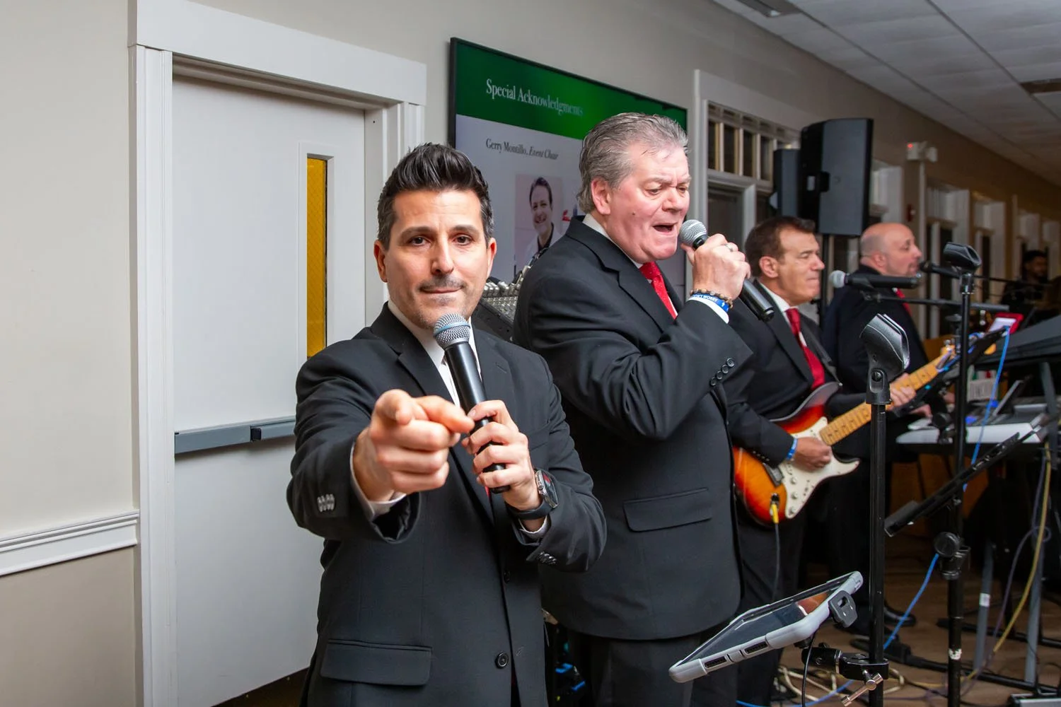Group of men in suits performing with microphones and guitar at an indoor event.
