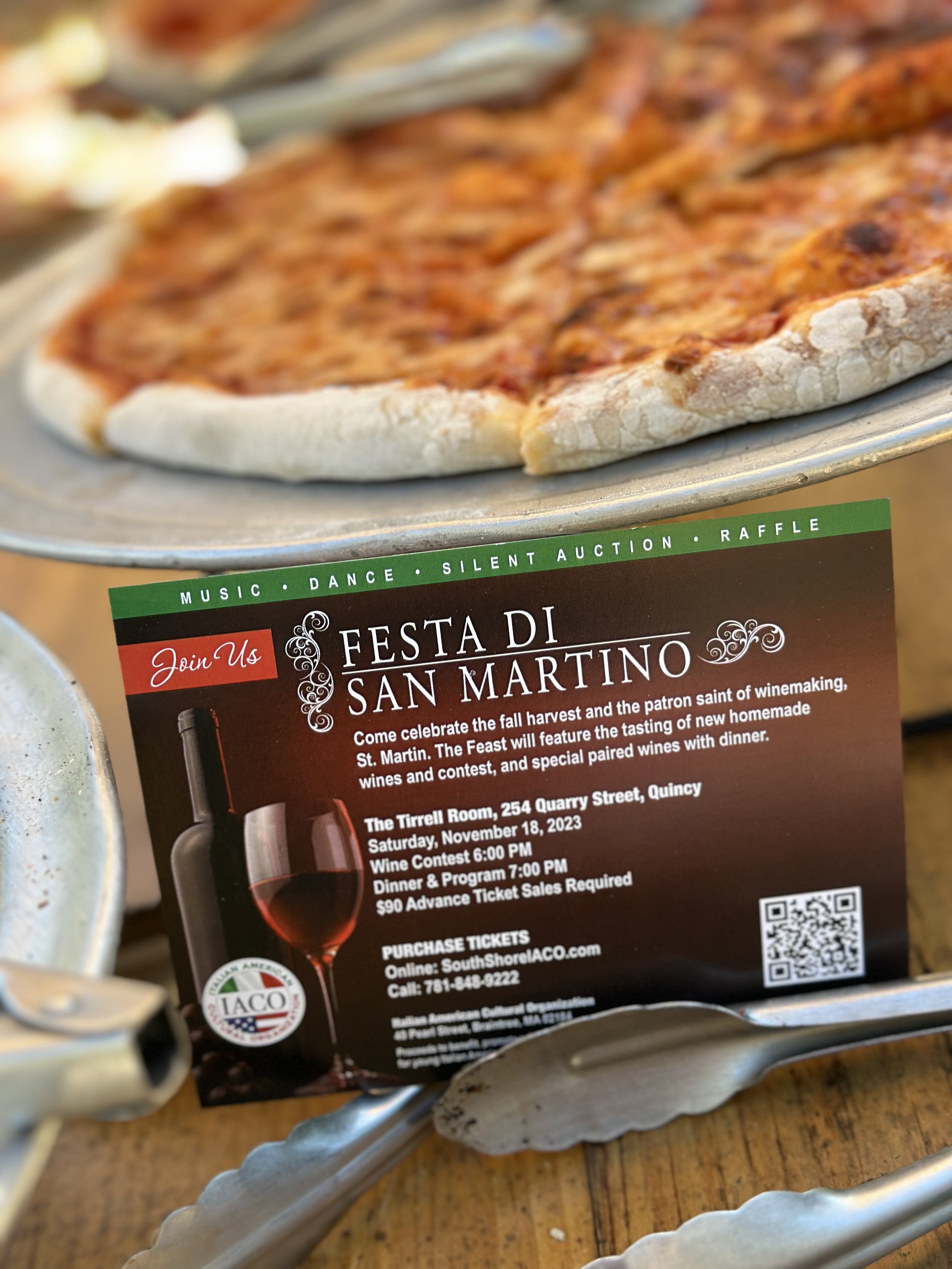 Close-up of a pepperoni pizza on a metal tray with a promotional card for Festa di San Martino, featuring event details and a bottle of wine and glass.