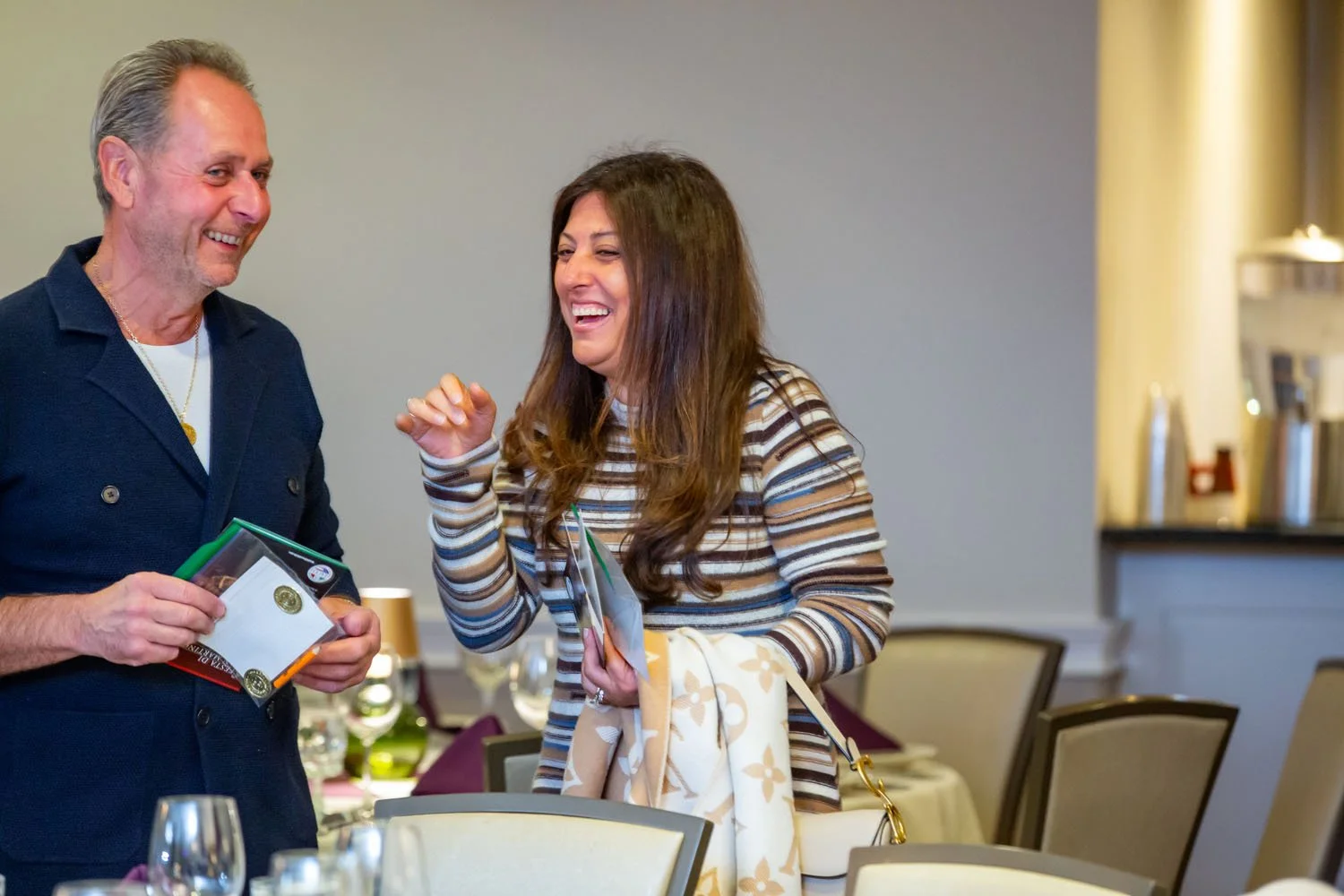 Two people smiling and talking in a dining room, one woman holding a small gift bag and a man holding a boxed item, with tables and chairs in the background.