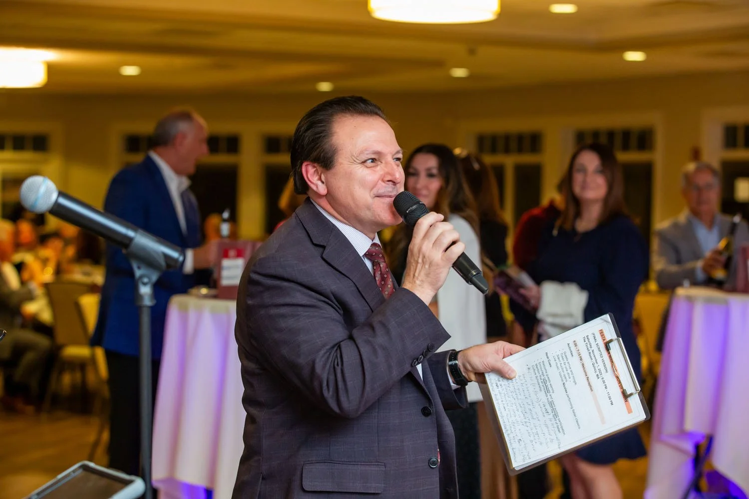 Man in suit speaking into a microphone at a formal event, holding a clipboard, with other people in the background at tables.