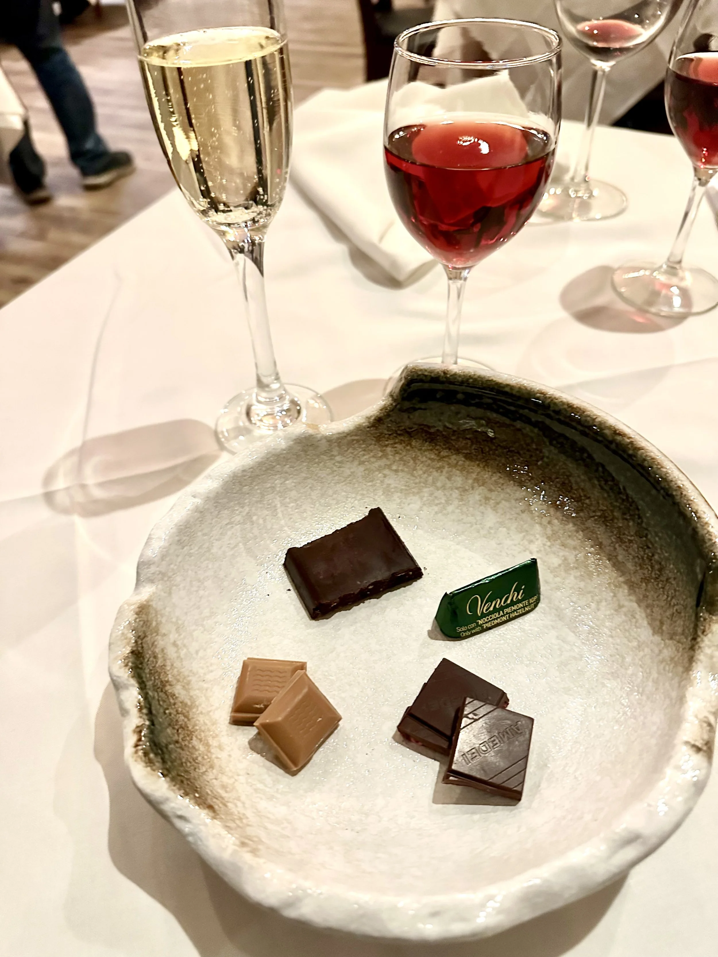A white ceramic bowl with five pieces of assorted chocolates on a white tablecloth, with four glasses of wine and a champagne flute in the background.