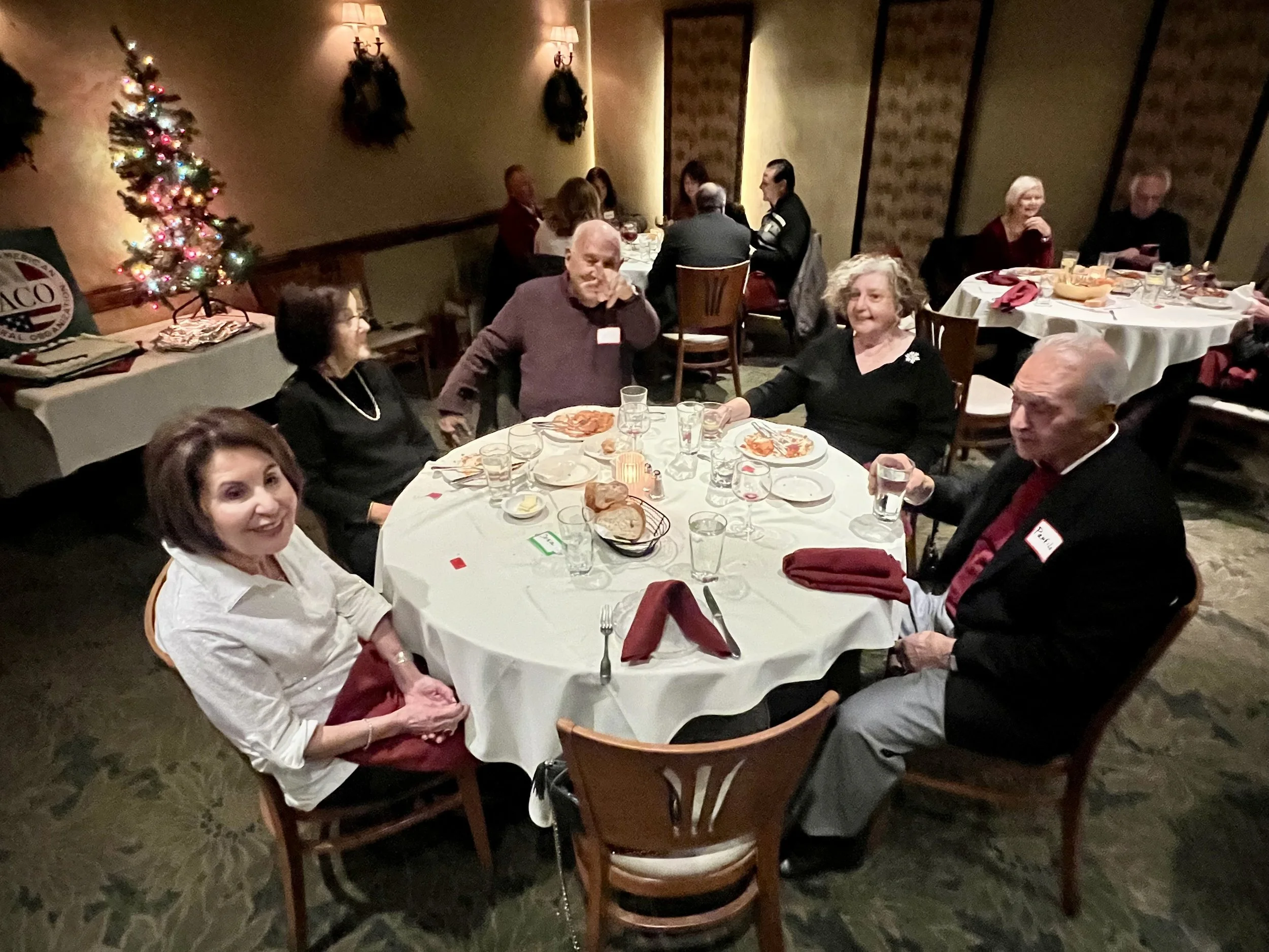 People sitting around a Holiday dinner table with Christmas decorations, including a lit Christmas tree, in a festive restaurant setting.