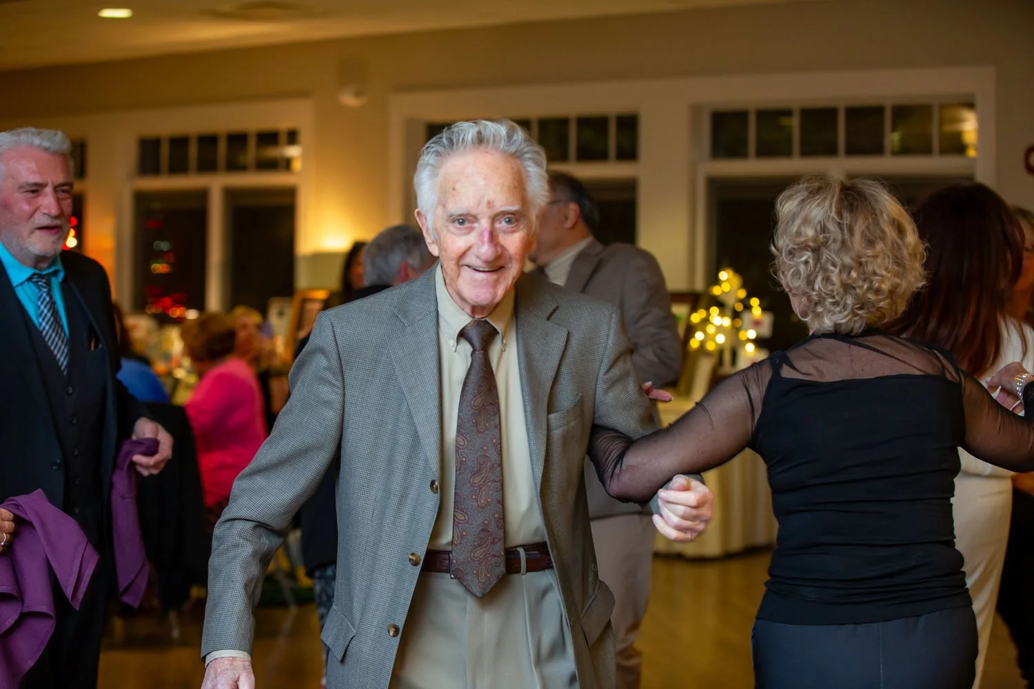 Older man in gray suit and beige tie dancing with woman in black dress with sheer sleeves at a festive gathering.