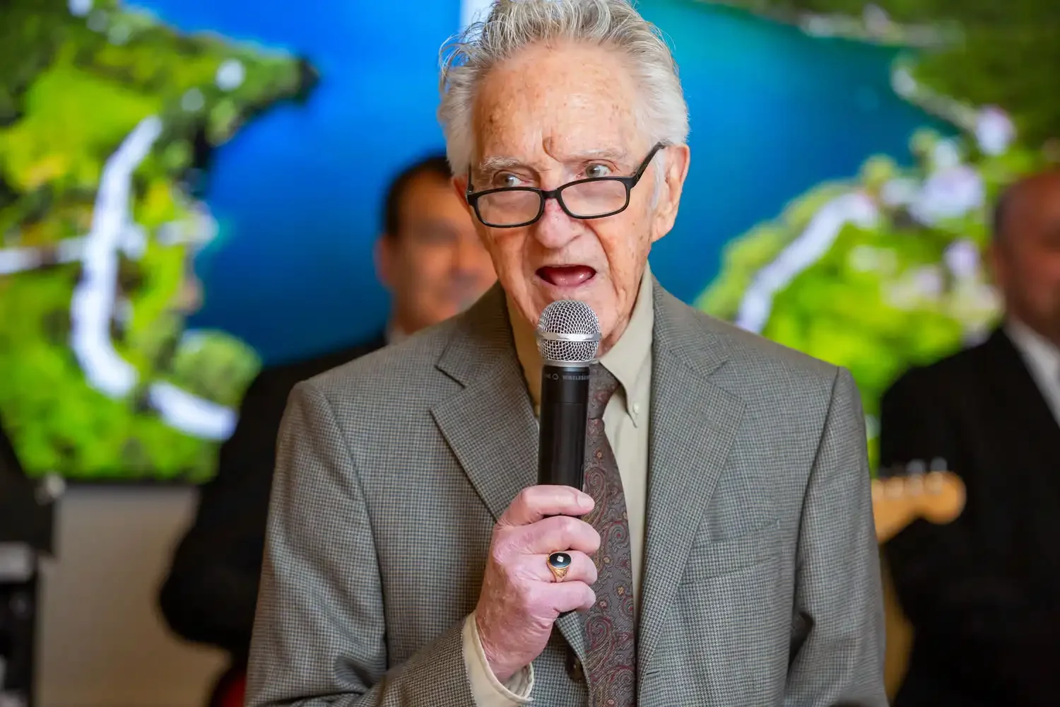 An elderly man with glasses, wearing a suit, speaking into a microphone at an event with a colorful backdrop and other people present.