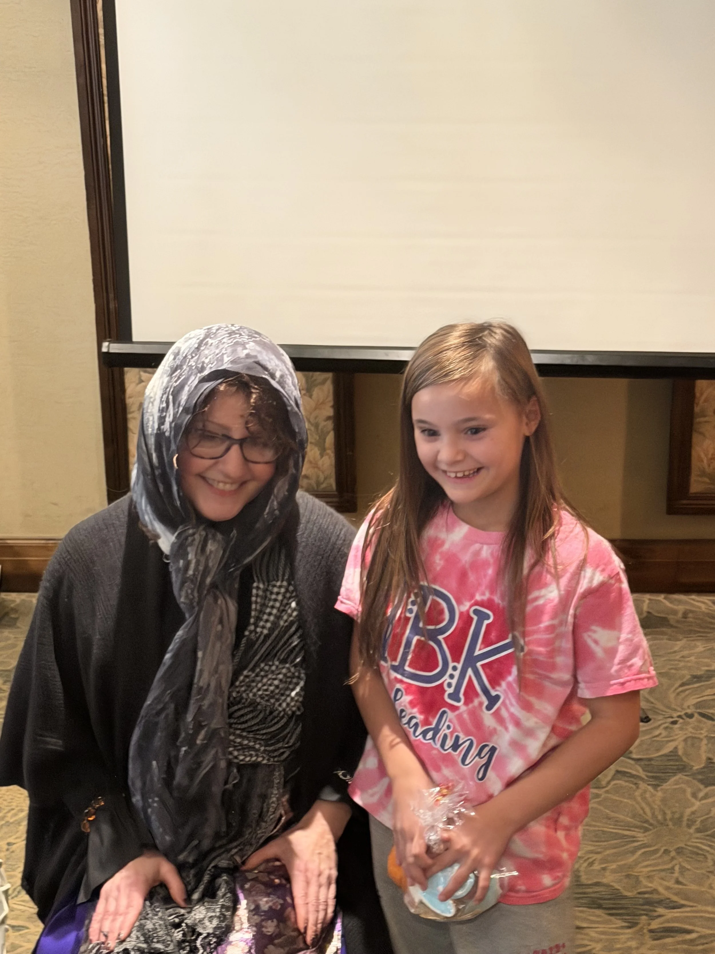 A woman with glasses and a patterned headscarf smiling next to a girl in a pink tie-dye T-shirt, holding a wrapped gift.