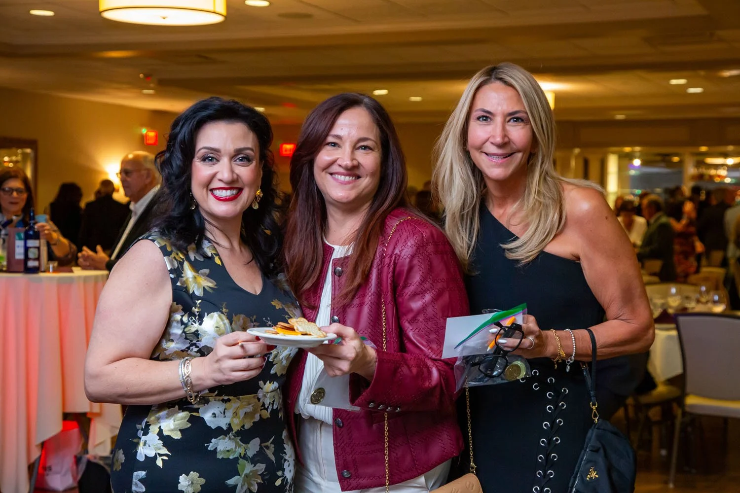 Three women smiling at a social event, standing close together. The woman on the left has dark hair, wearing a floral dress, holding a small plate. The woman in the middle has reddish-brown hair, wearing a red jacket. The woman on the right has blond
