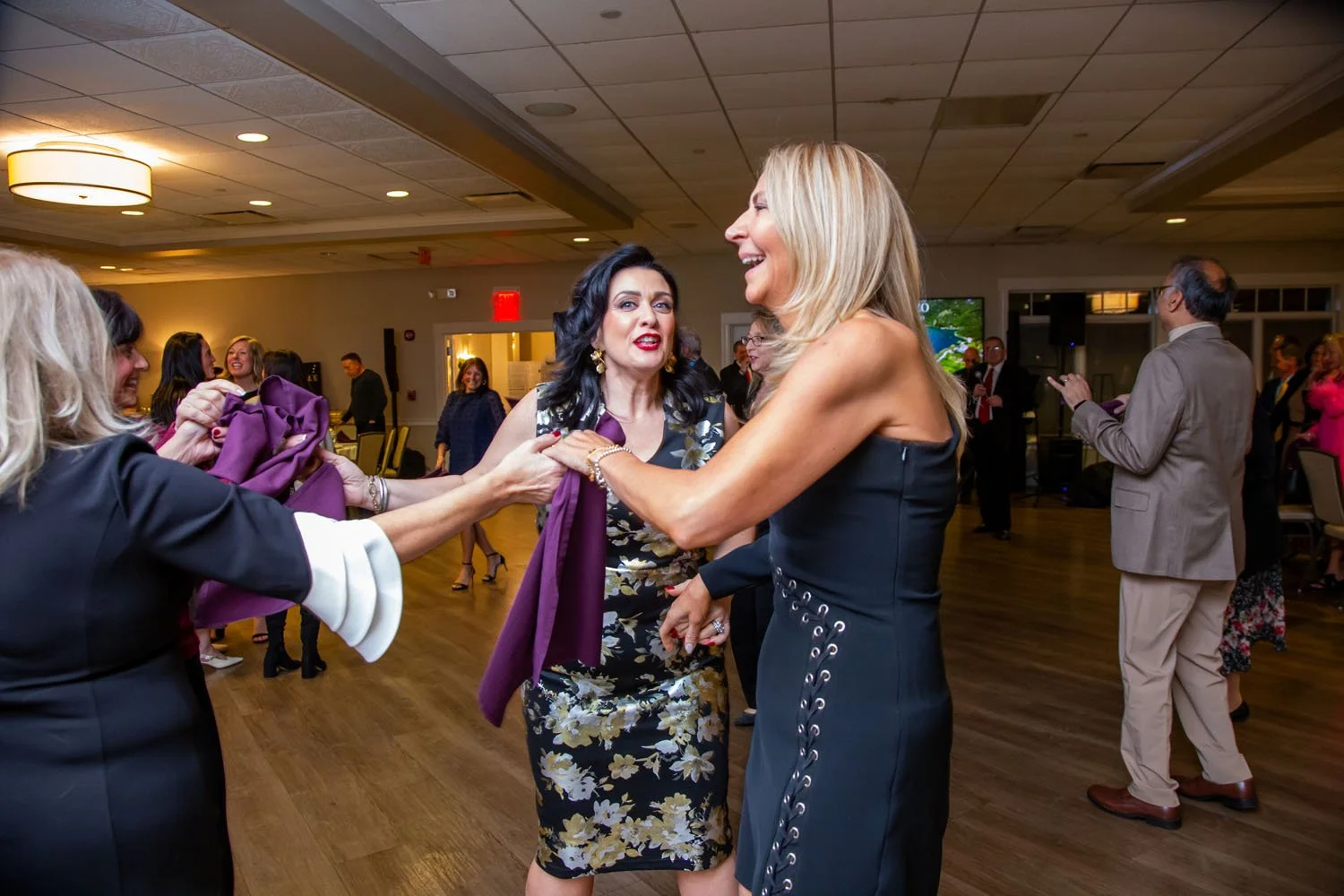 Two women dancing and laughing at a social event, with other people in the background on a wooden floor, in a well-lit room.