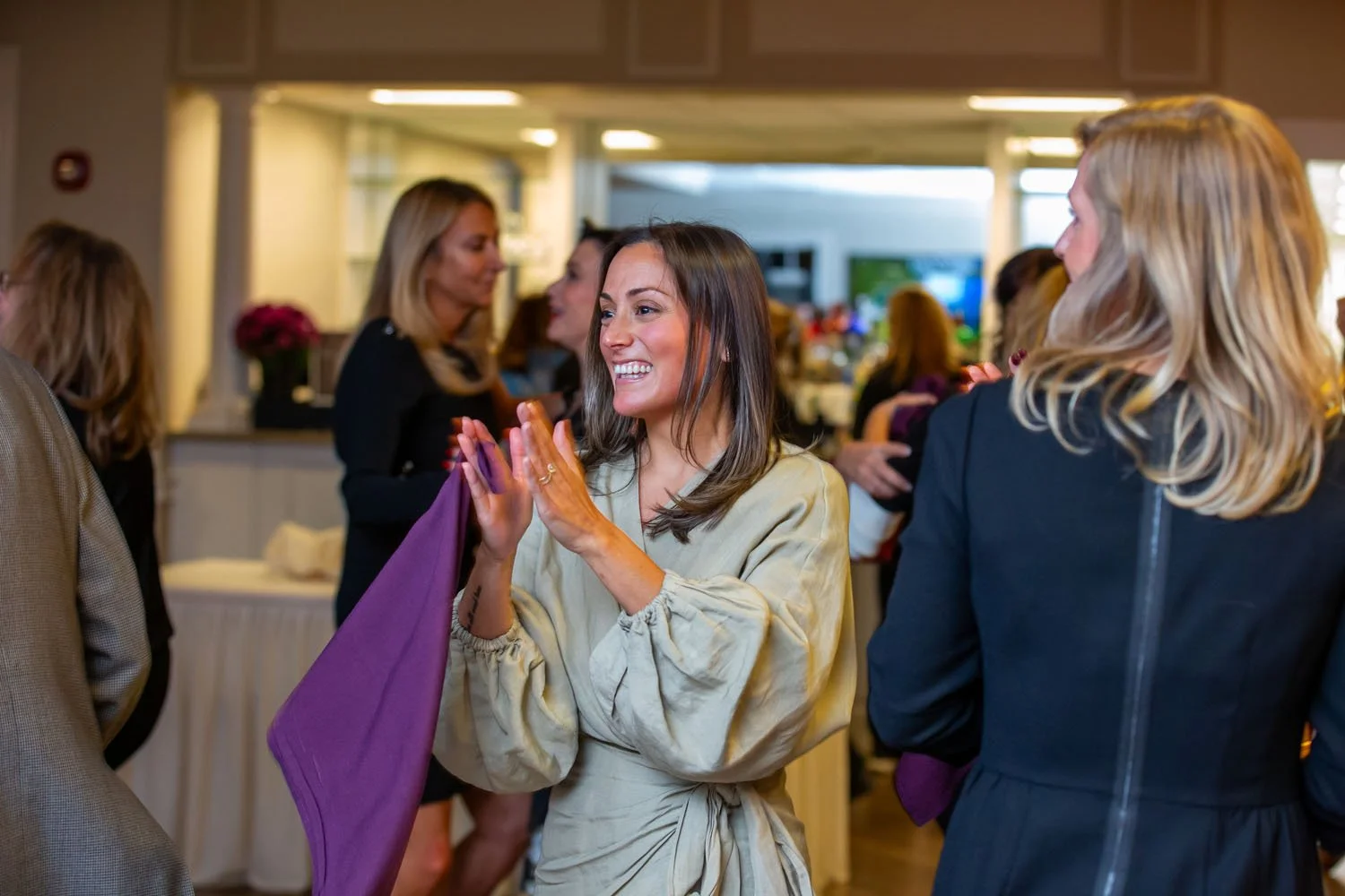 A woman in a beige dress is smiling and clapping her hands in a social gathering, surrounded by other women in a well-lit indoor space.