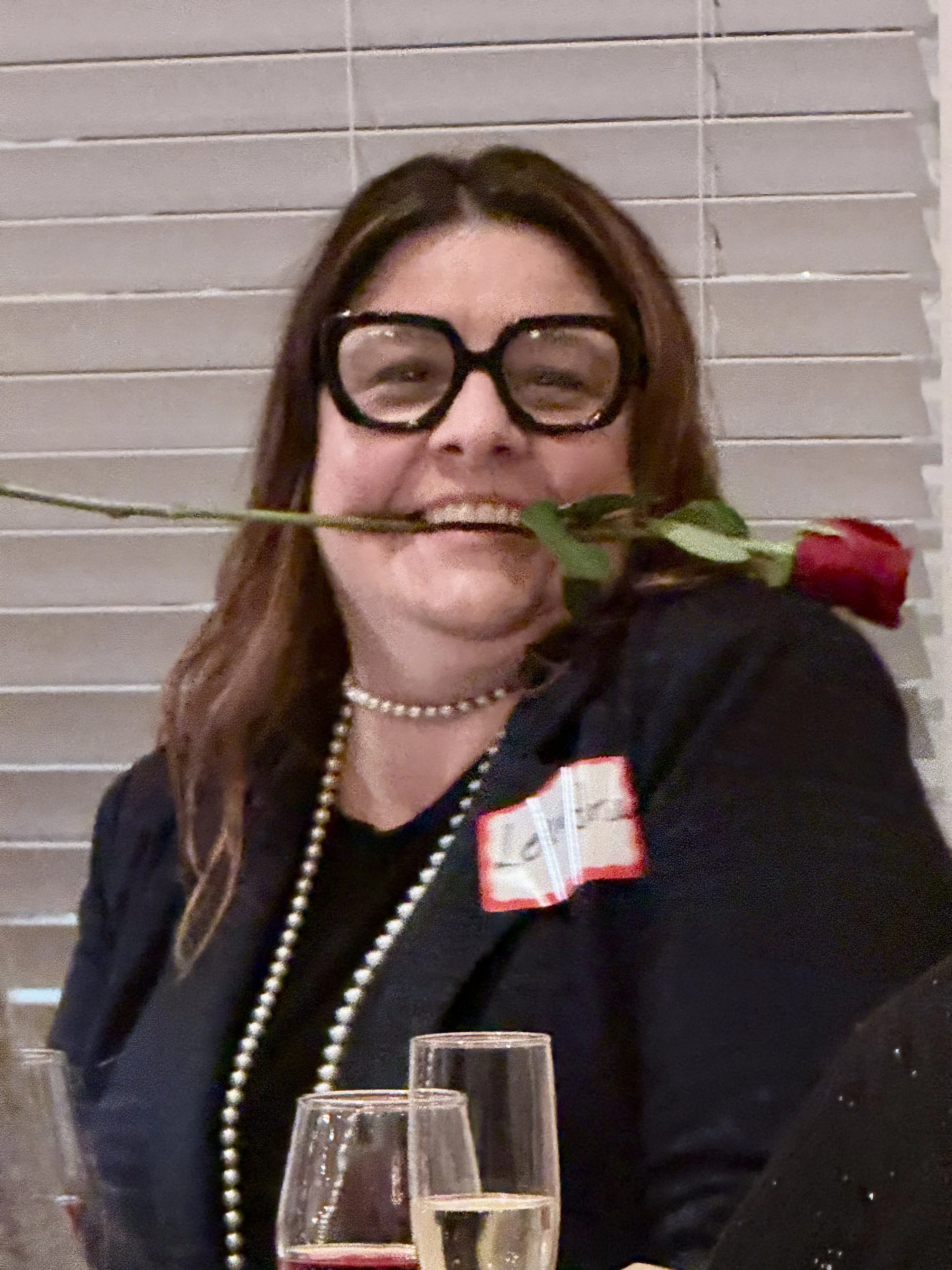 Woman with long brown hair wearing large black glasses, pearl necklace, and a black jacket, holding a single red rose in her mouth at a social gathering. There are wine glasses in front of her and a background with horizontal blinds.