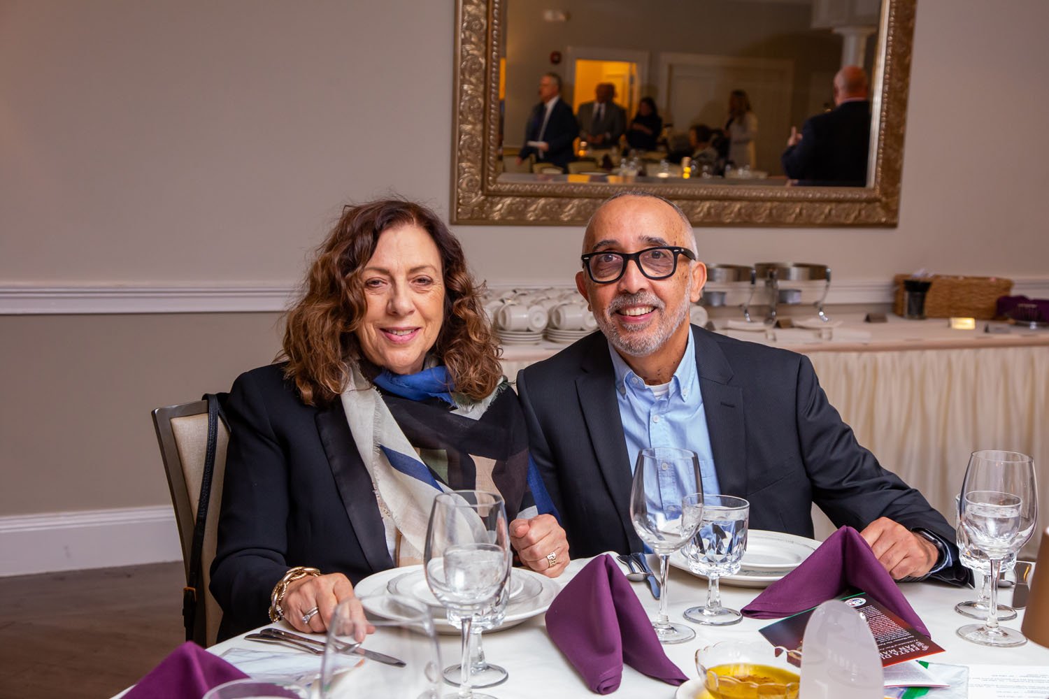 A smiling older woman with curly brown hair and an older man with glasses and a beard, dressed in formal attire, sitting at a dinner table with purple napkins, wine glasses, and plates, in a well-lit restaurant or banquet hall. They are seated side b