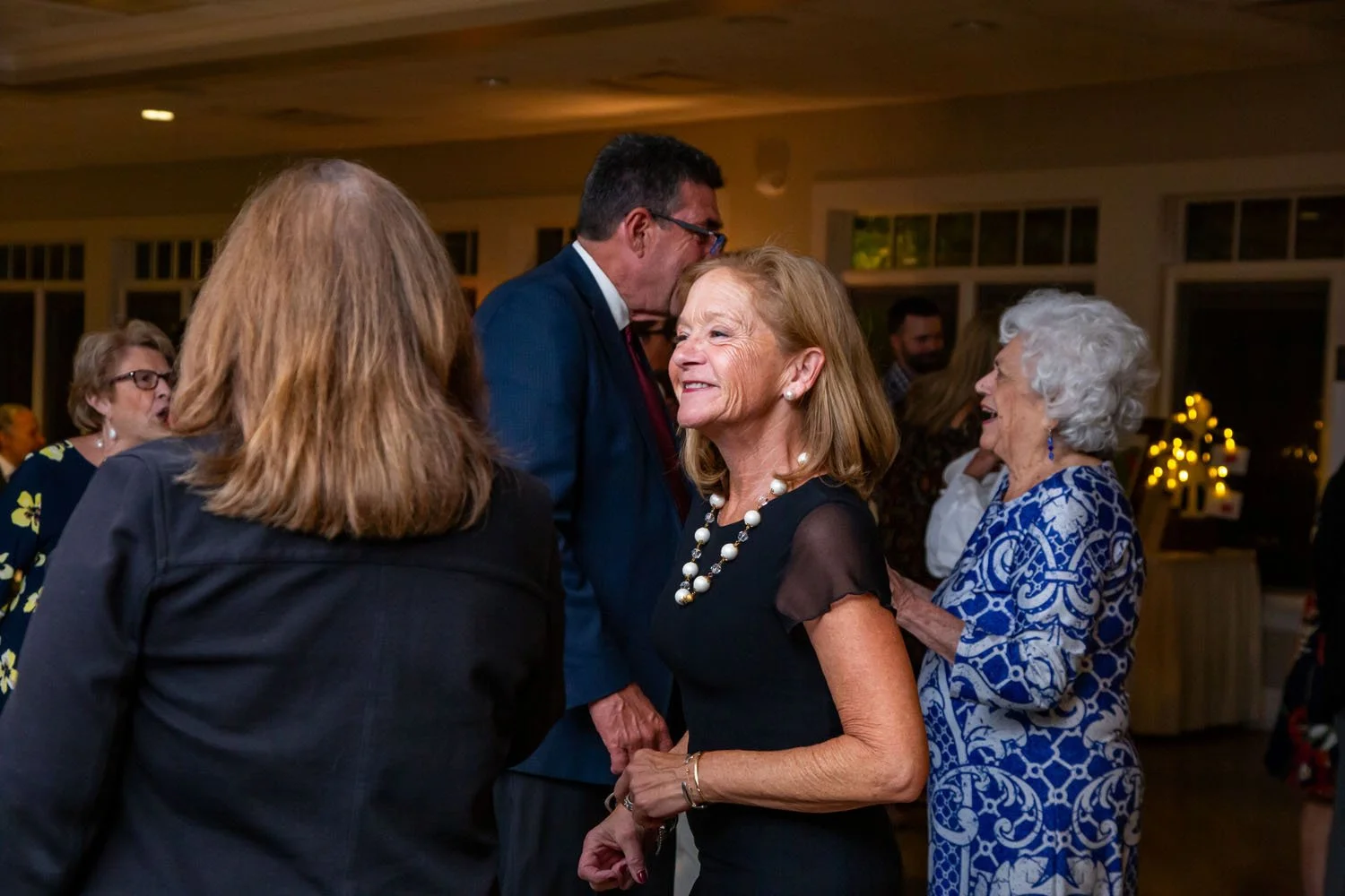 A group of mature adults at a social gathering, with a woman smiling and conversing with a man. Others are talking in the background, in a warmly lit room with windows and decorative lighting.
