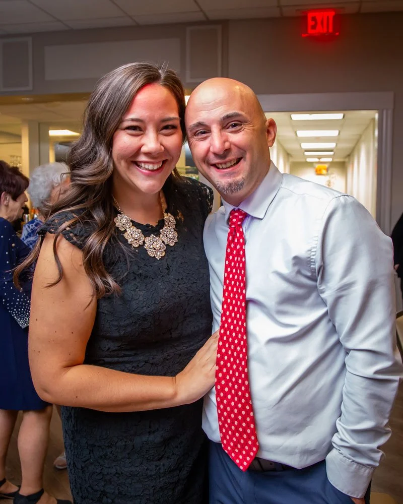 A smiling woman in a black lace dress with a statement necklace, posing with a smiling man in a white shirt and red polka dot tie, indoors at a social event.