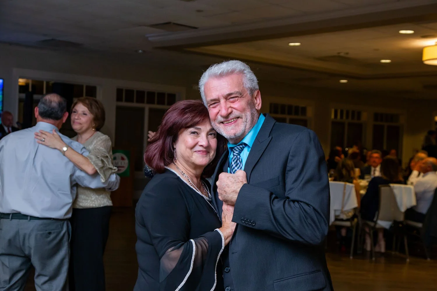 A smiling elderly couple dancing at a formal event, with other dancing couples and seated guests in the background.