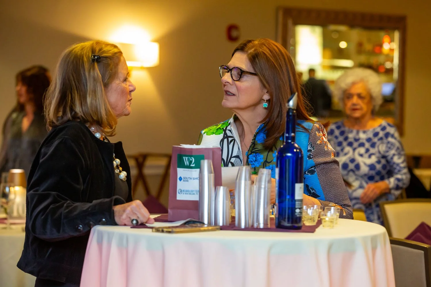 Two older women having a conversation at a round table in a restaurant or event space, with another woman in the background watching, in a warm lit, social setting.