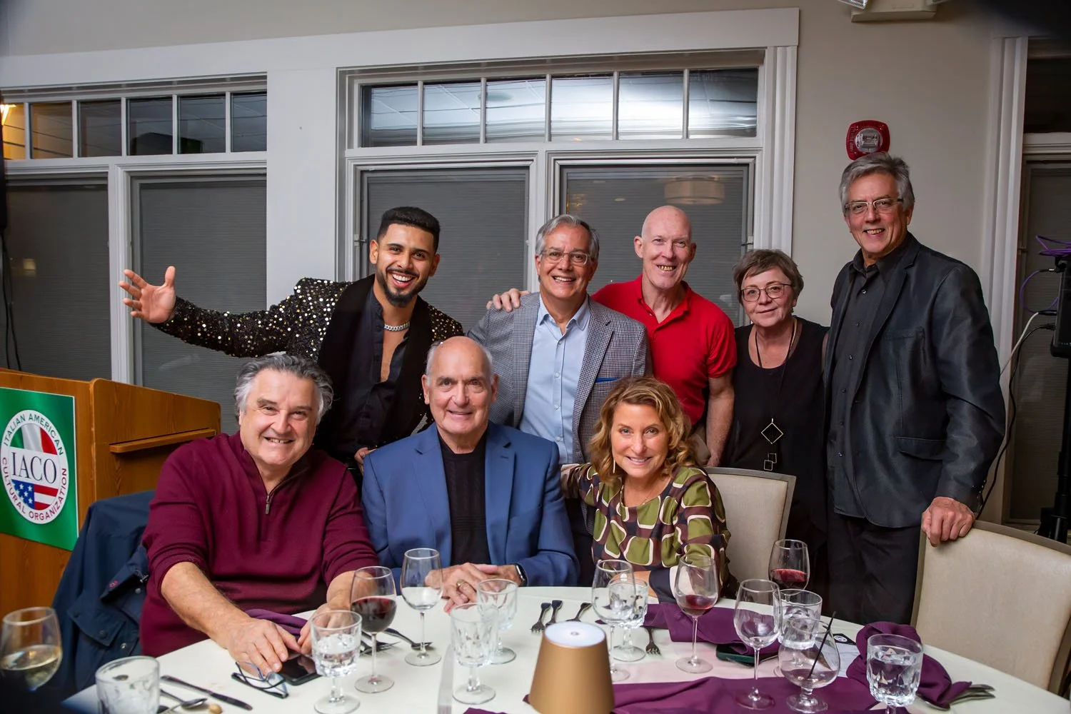 Group of nine people gathered around a dining table with wine glasses, smiling for a photo indoors at a formal event.