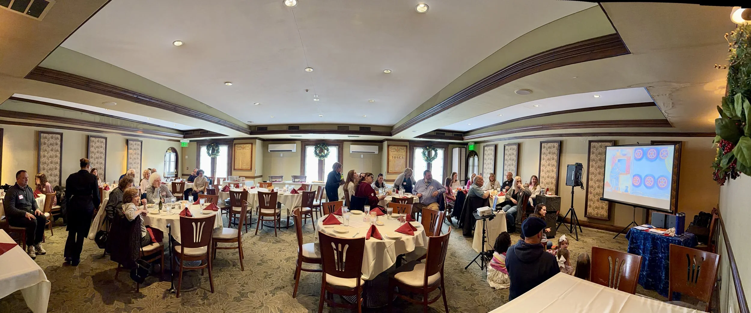 People gathered in a decorated banquet hall watching a presentation or performance on a television screen, with tables set for a meal and holiday wreaths hanging in the windows.
