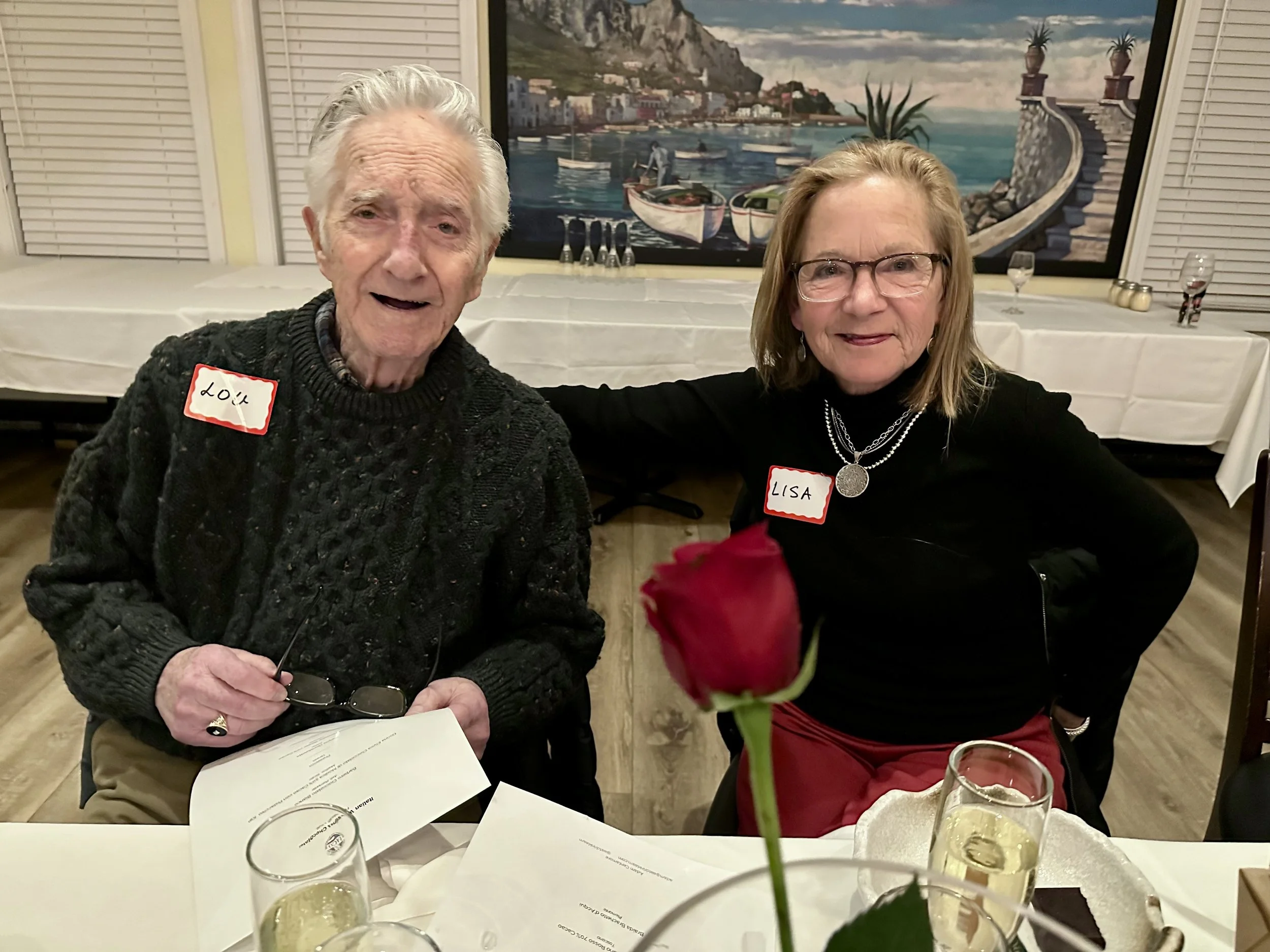 Older man and woman sitting at a table with white tablecloth, smiling at the camera, with a red rose in the foreground, in a decorated restaurant with a large mural of a coastal scene in the background.