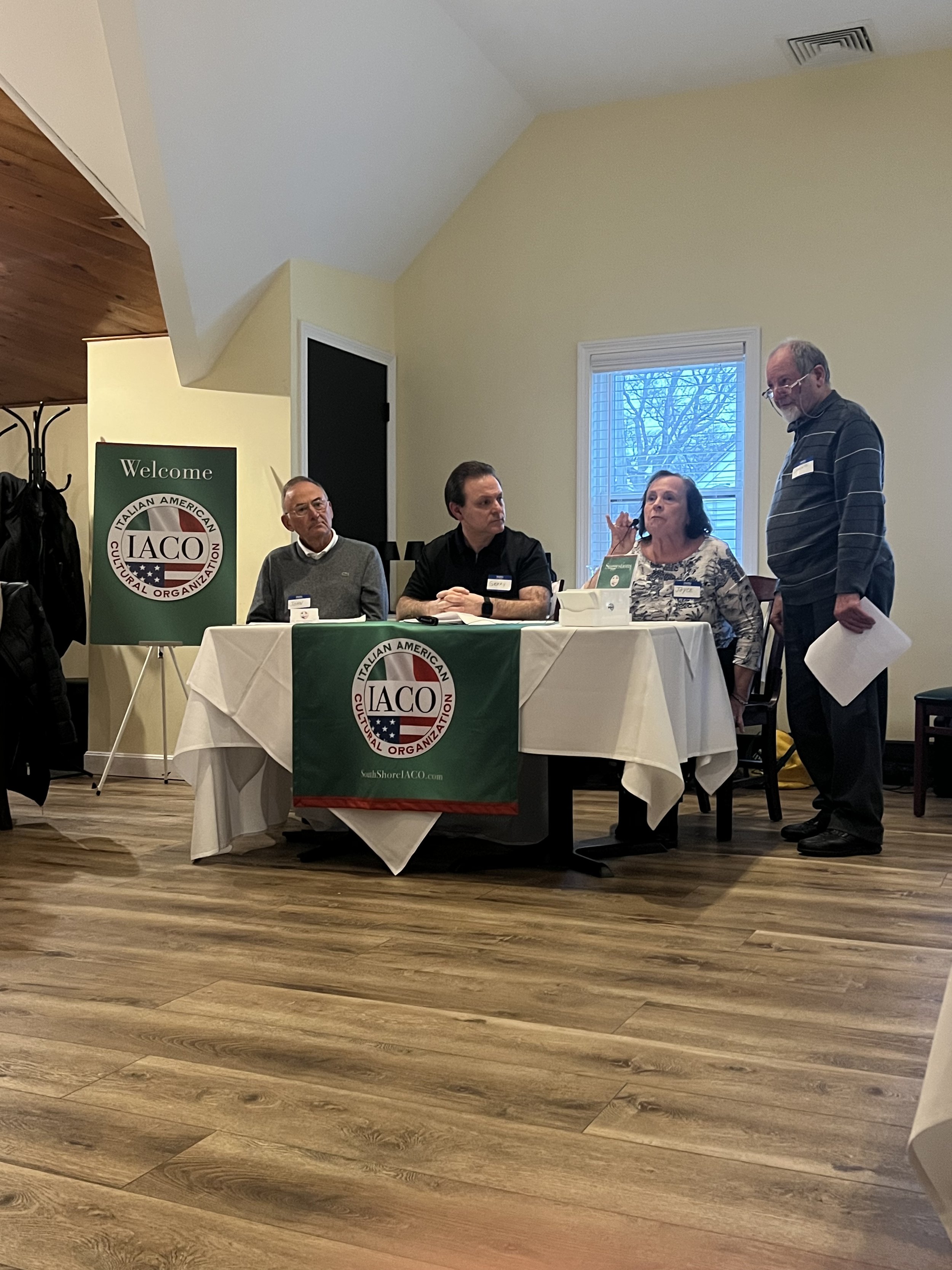 Four people sitting and standing around a table with a banner for the Italian American Cultural Organization (IACO). One person is speaking while others listen during a meeting or event in a room with wooden flooring and a welcome sign displayed.