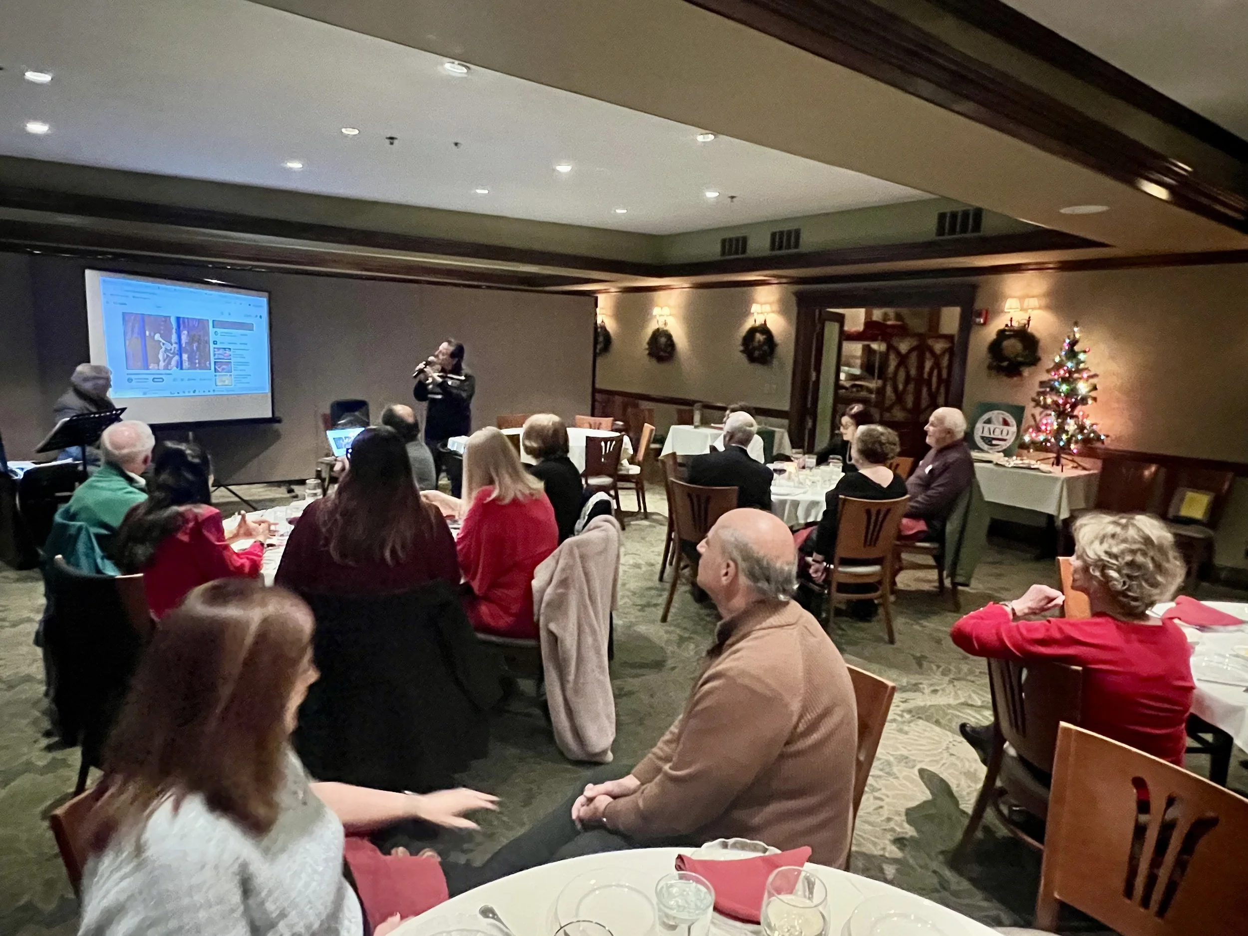 People sitting at tables in a dimly lit banquet room watching a woman perform with a clarinet. There is a Christmas tree and holiday decorations in the background.