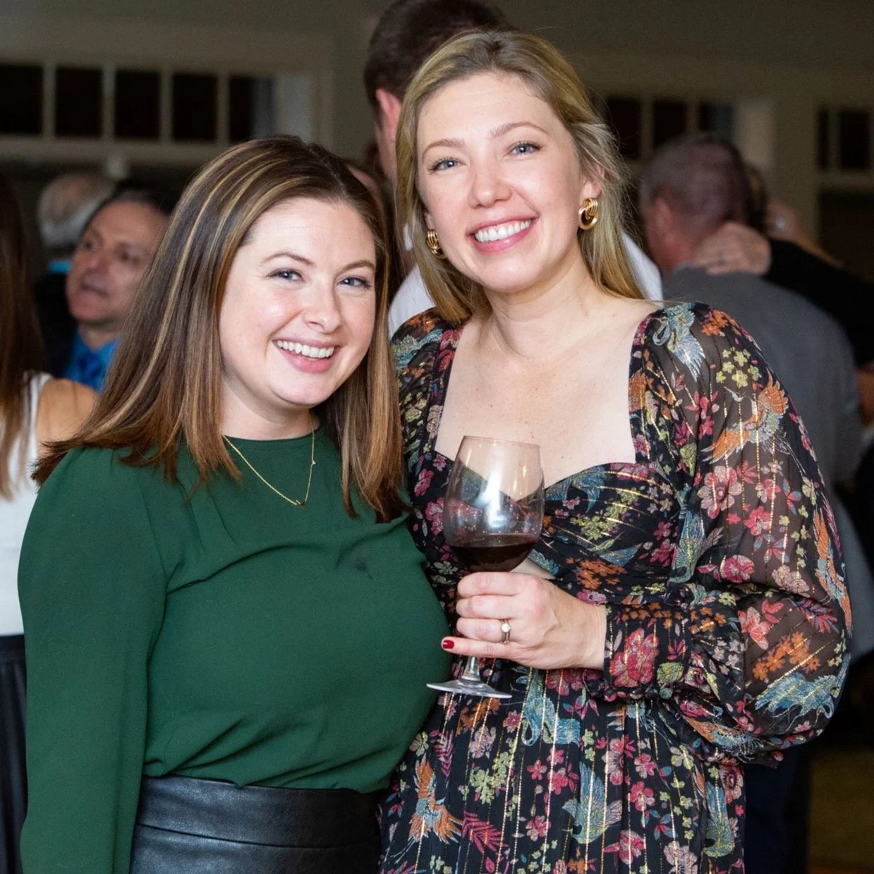 Two women smiling at a social gathering, one holding a glass of red wine, surrounded by other guests.